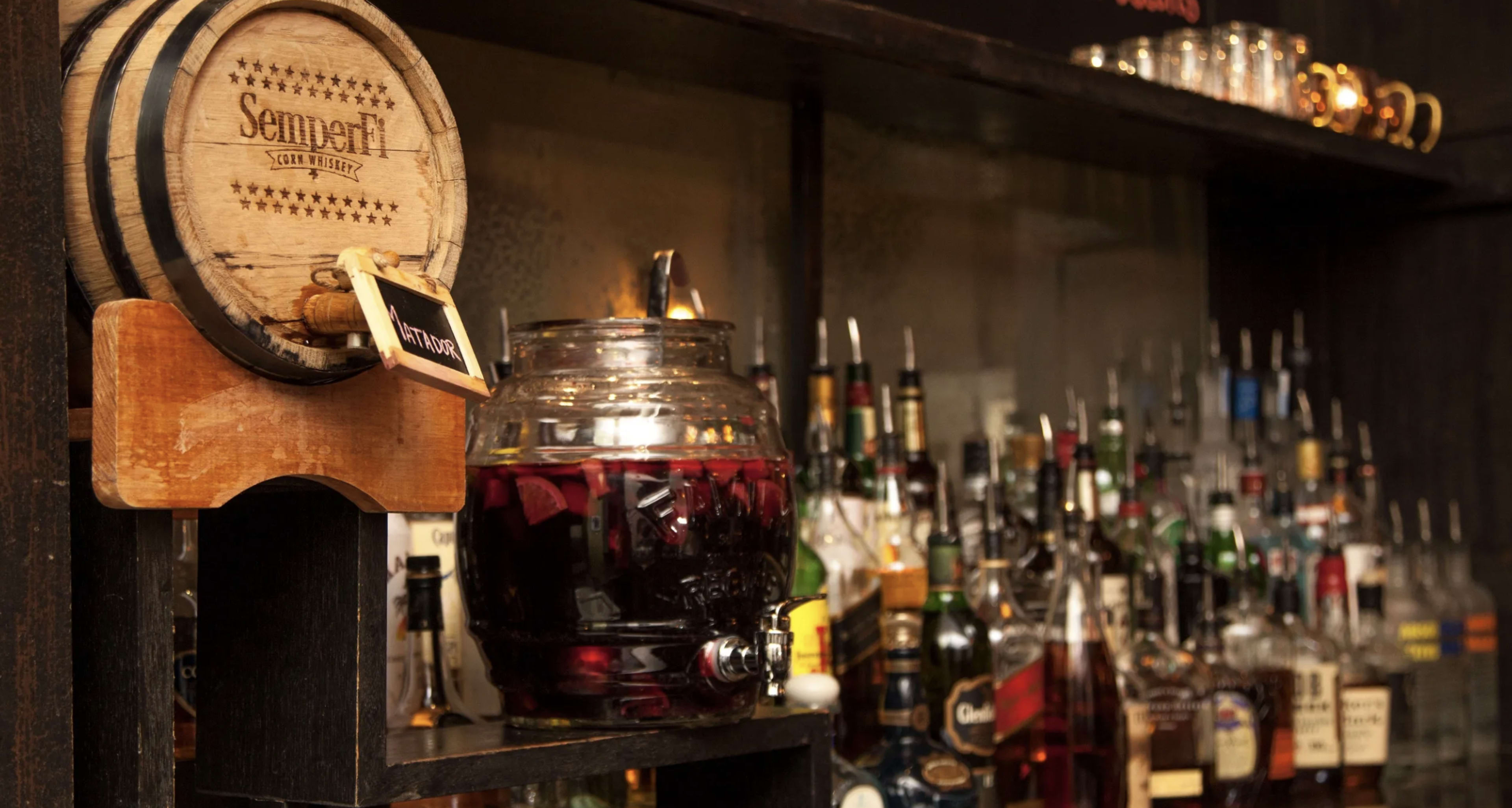 A wooden barrel with a label is displayed alongside a glass container filled with dark liquid, surrounded by various bottles of alcohol on a shelf.