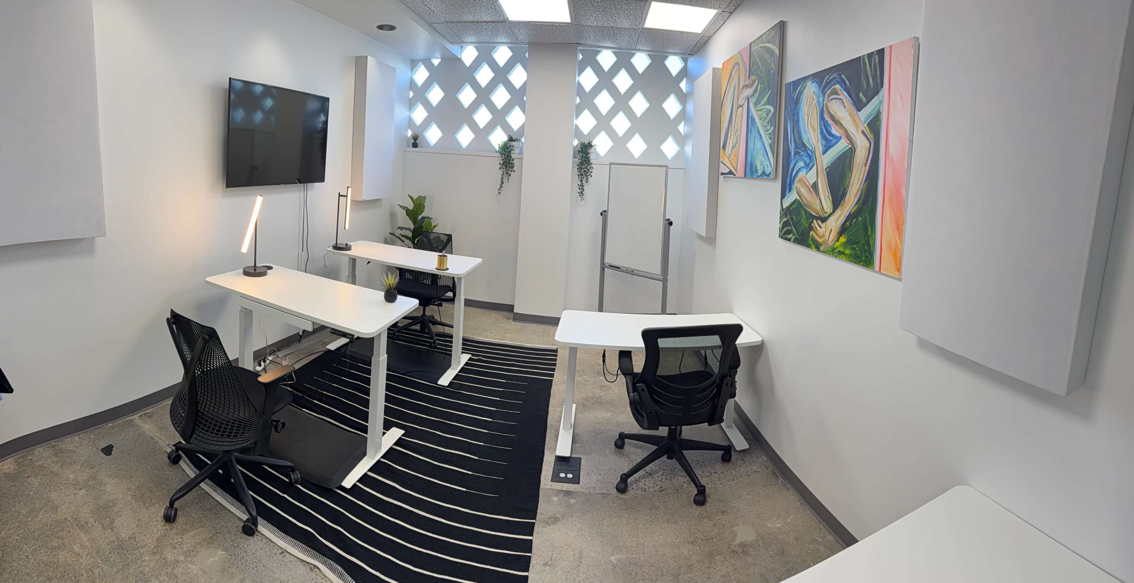 The image shows a modern office space with two desks, a television mounted on the wall, and colorful artwork displayed.