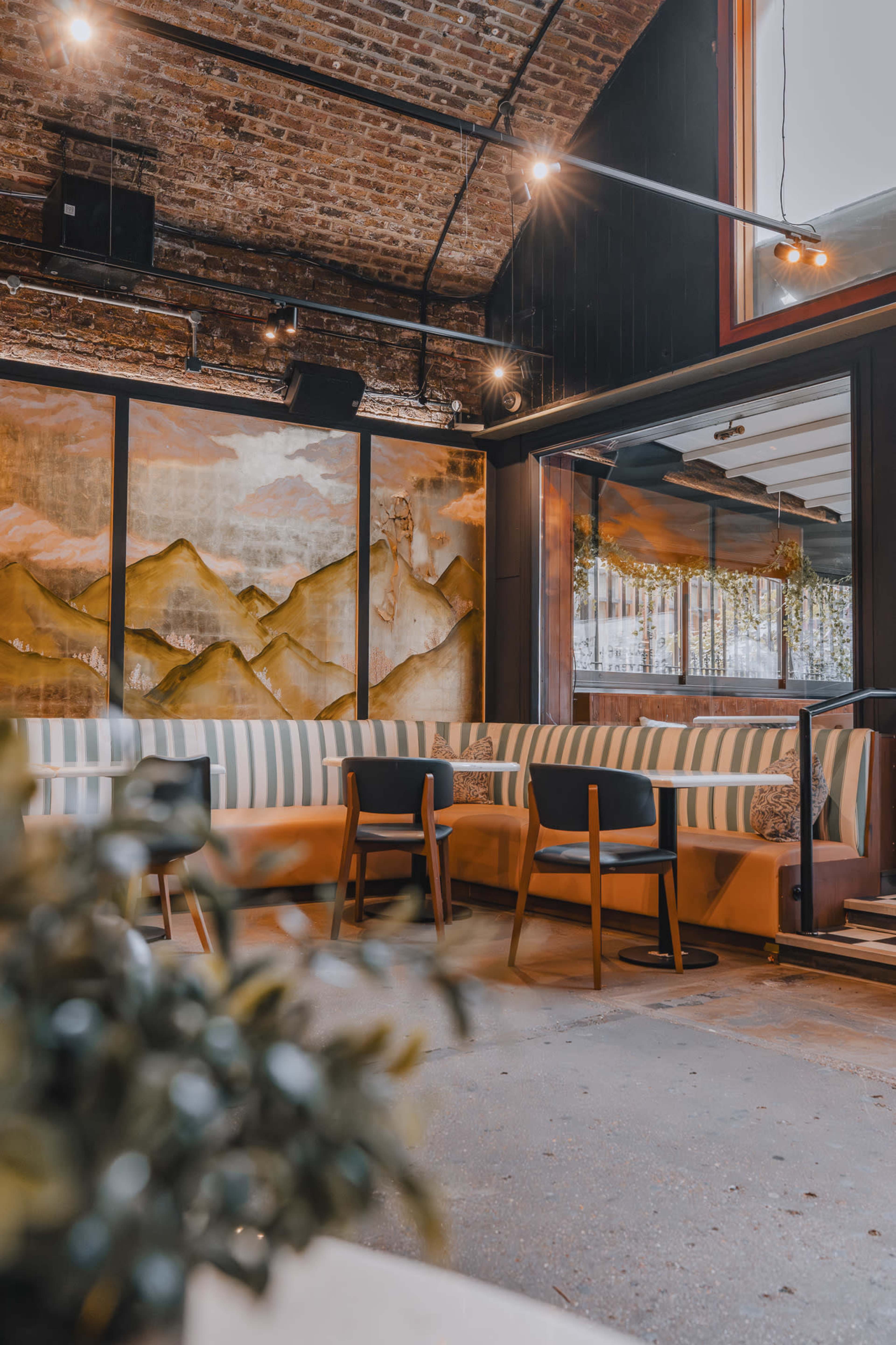 The image shows a cozy restaurant interior with a mix of striped seating, wooden chairs, and a large mountain mural on the wall.