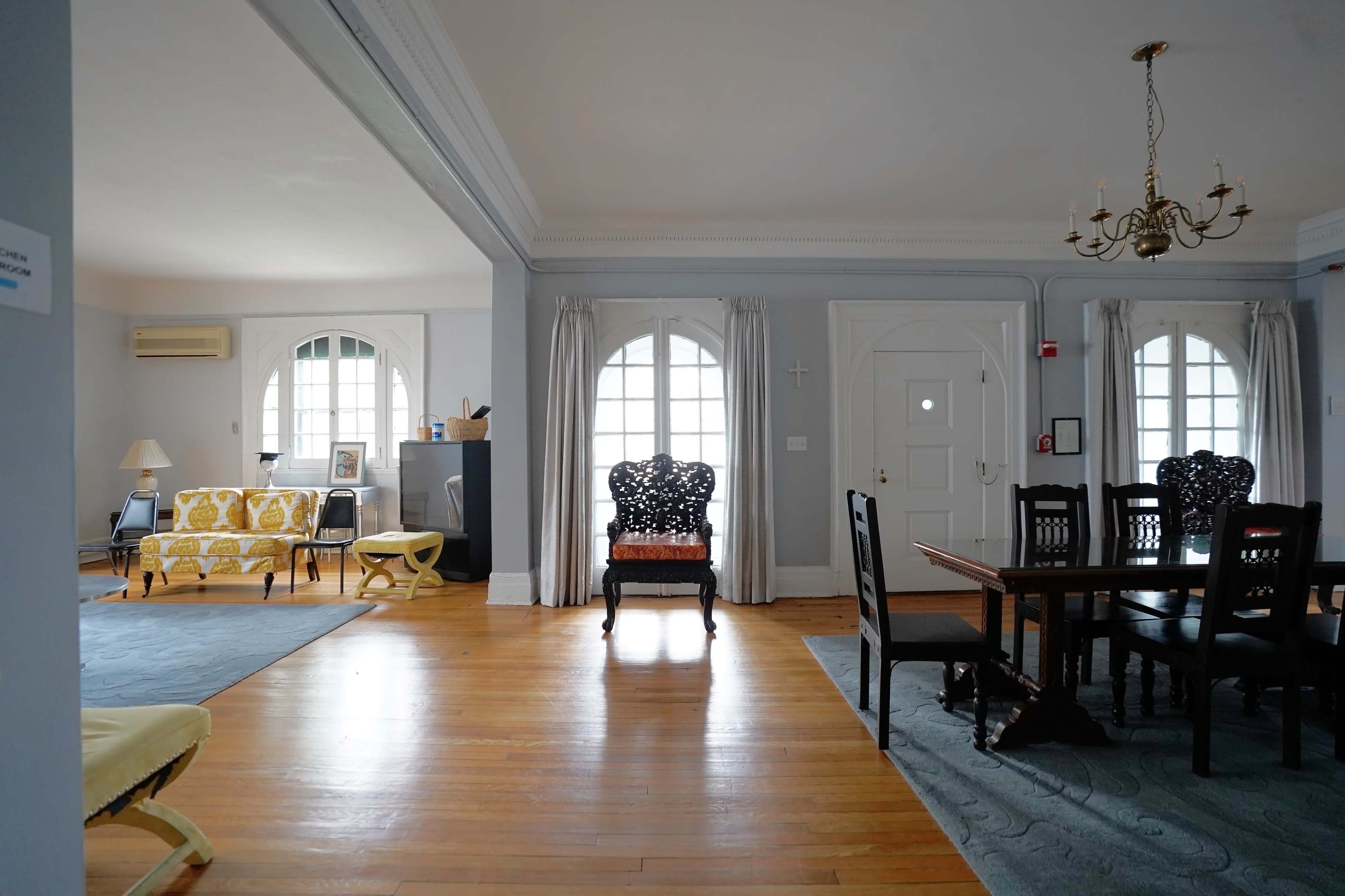 The image shows a spacious living area with wooden floors, featuring a yellow patterned couch, dark wooden dining chairs, and large arched windows.