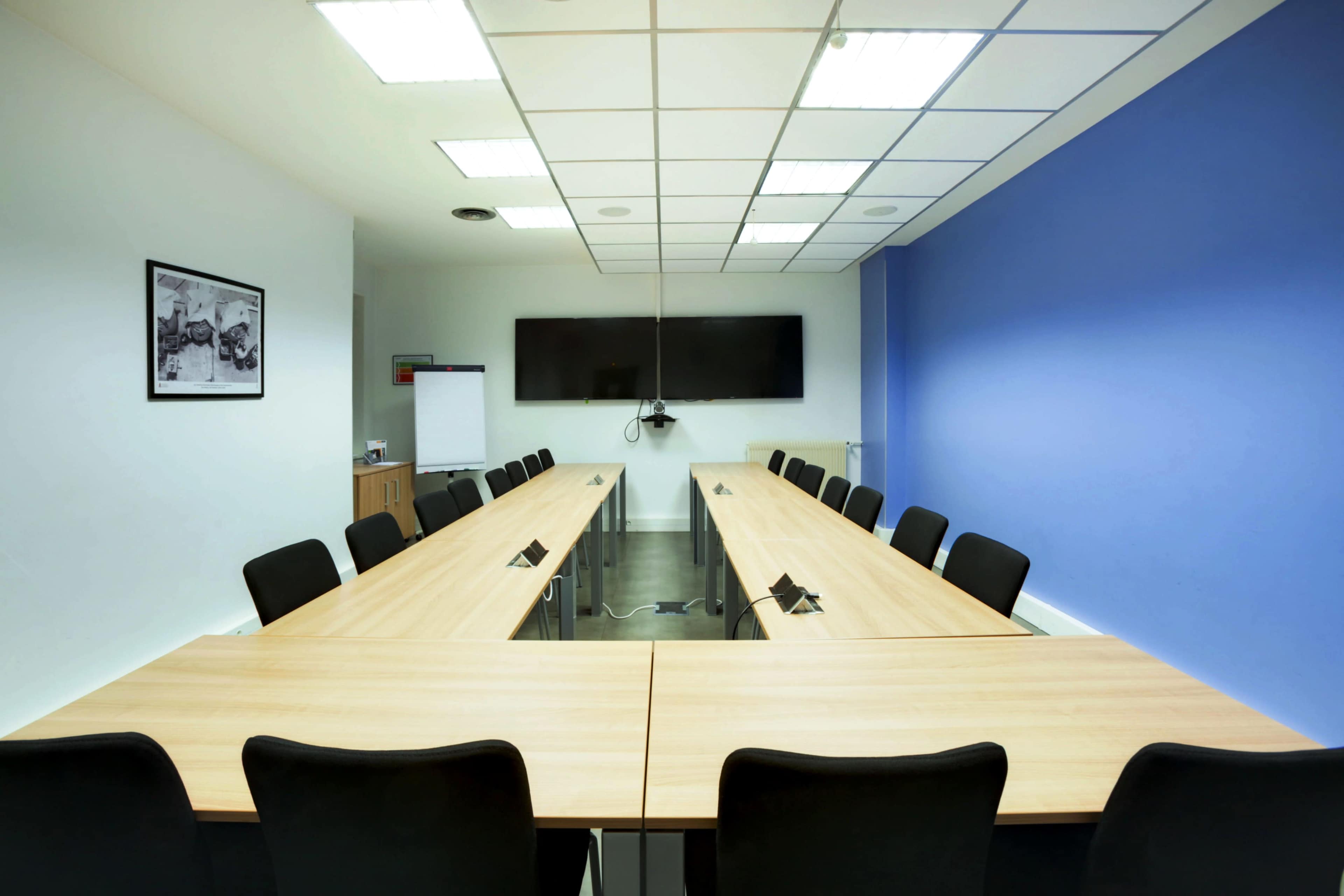 A conference room features two long wooden tables arranged in a U-shape, surrounded by black chairs, with a large screen mounted on the wall and a blue accent wall.