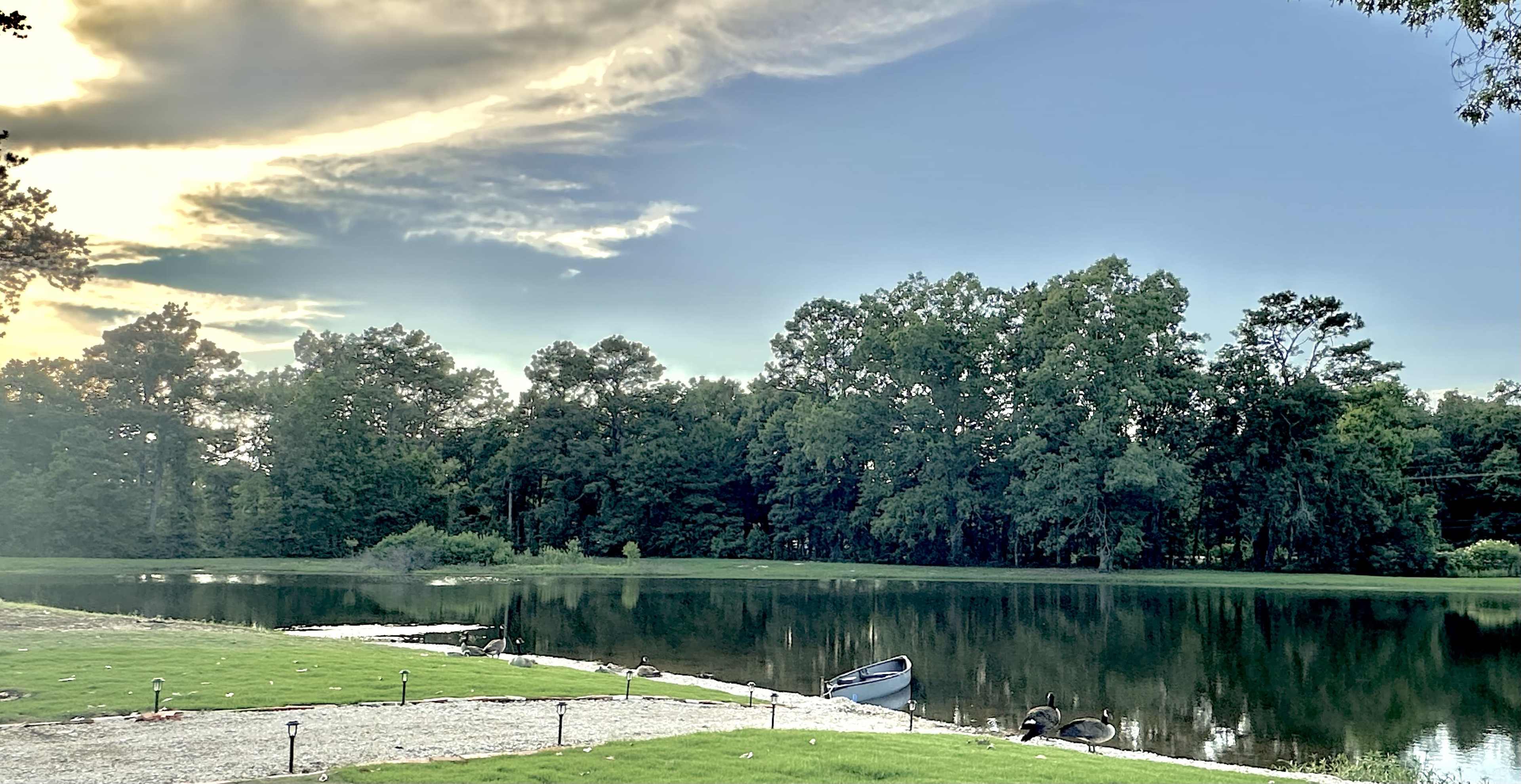 The image shows a calm lake surrounded by trees, with a small boat pulled up on the shore and landscaped grass leading to the water.