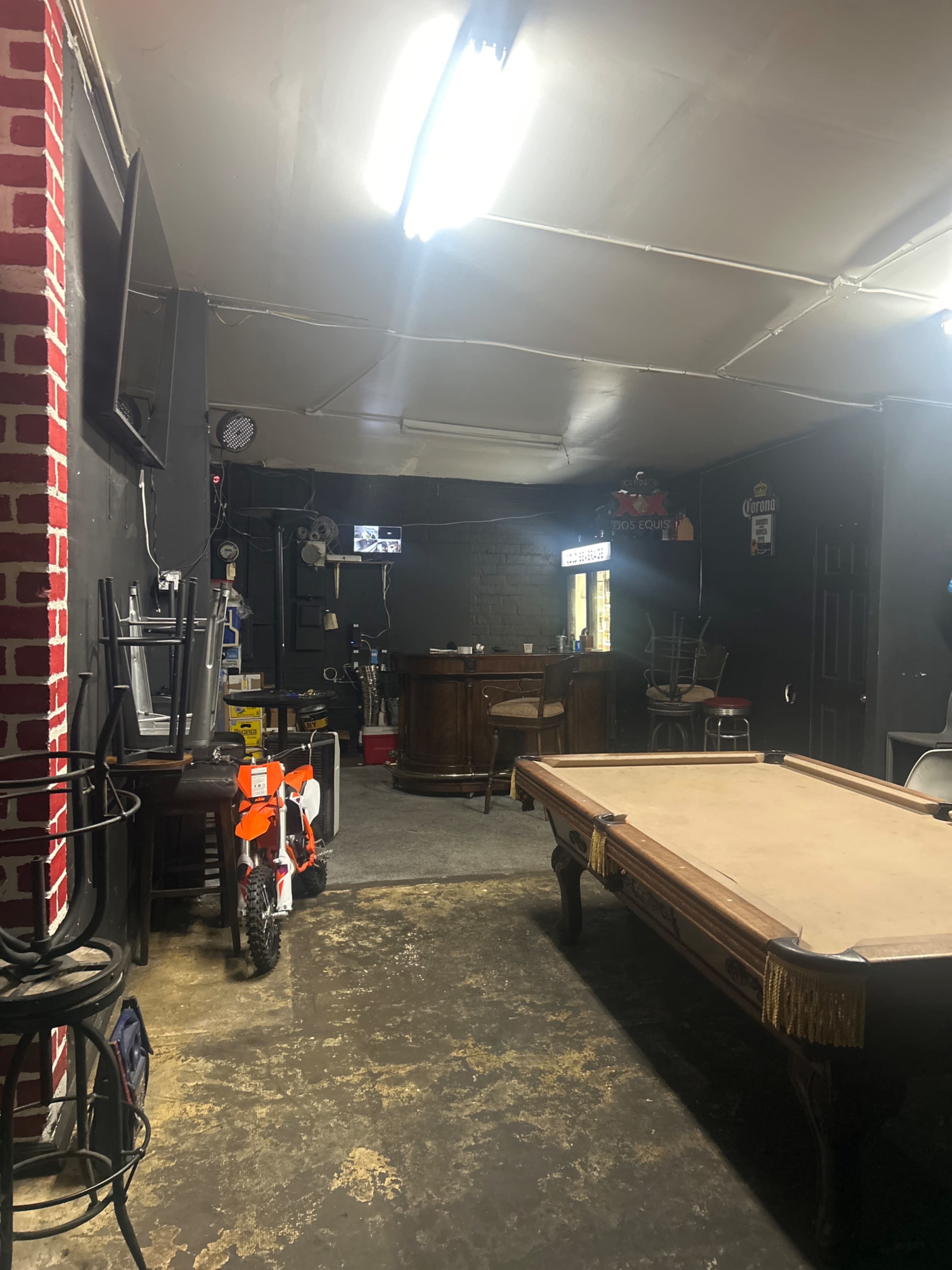 The image shows a dimly lit room featuring a pool table, a bar area, and a dirt bike leaning against the wall.