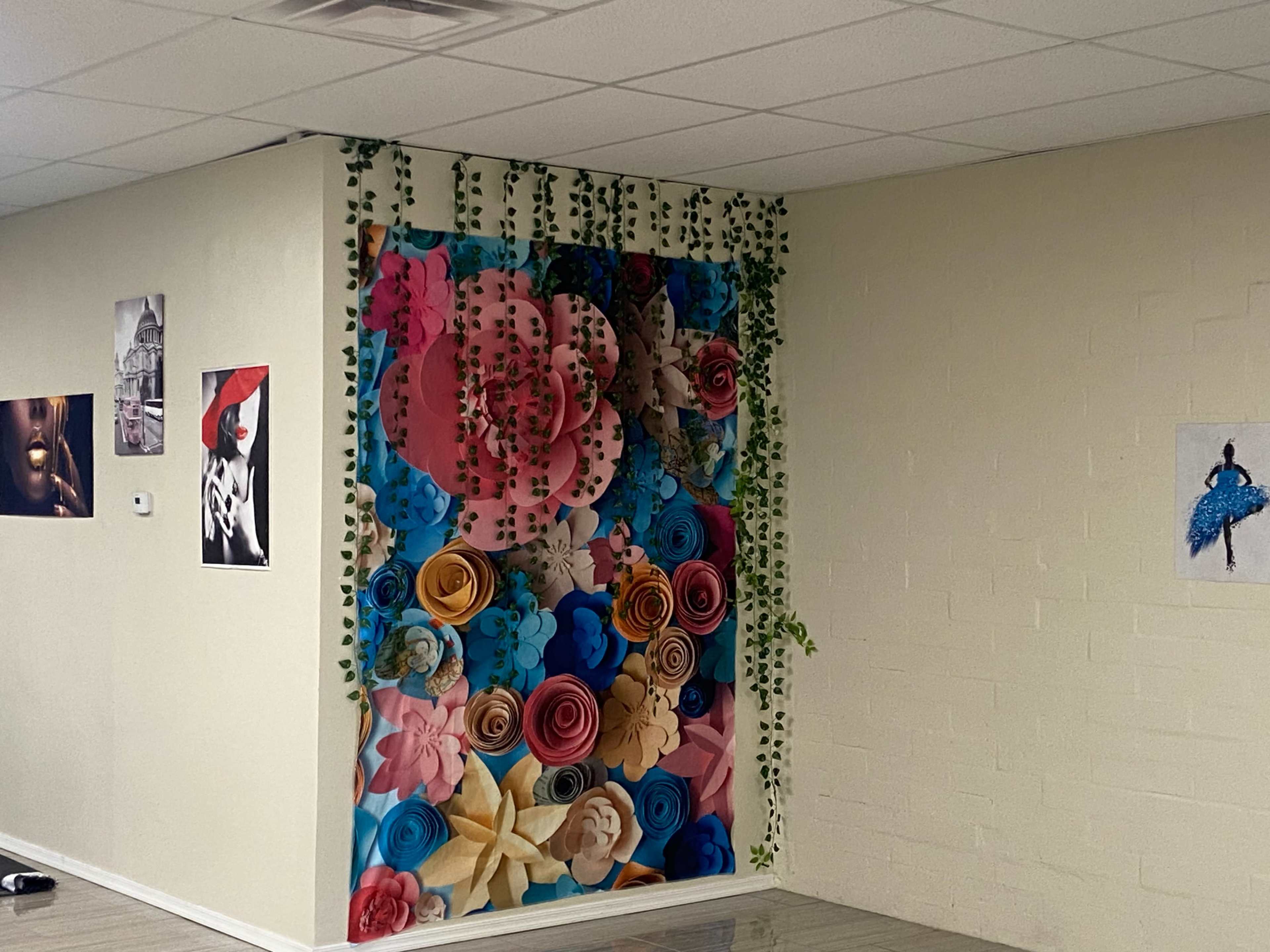 A wall decorated with large paper flowers in various colors, surrounded by green vines, alongside several framed artworks.