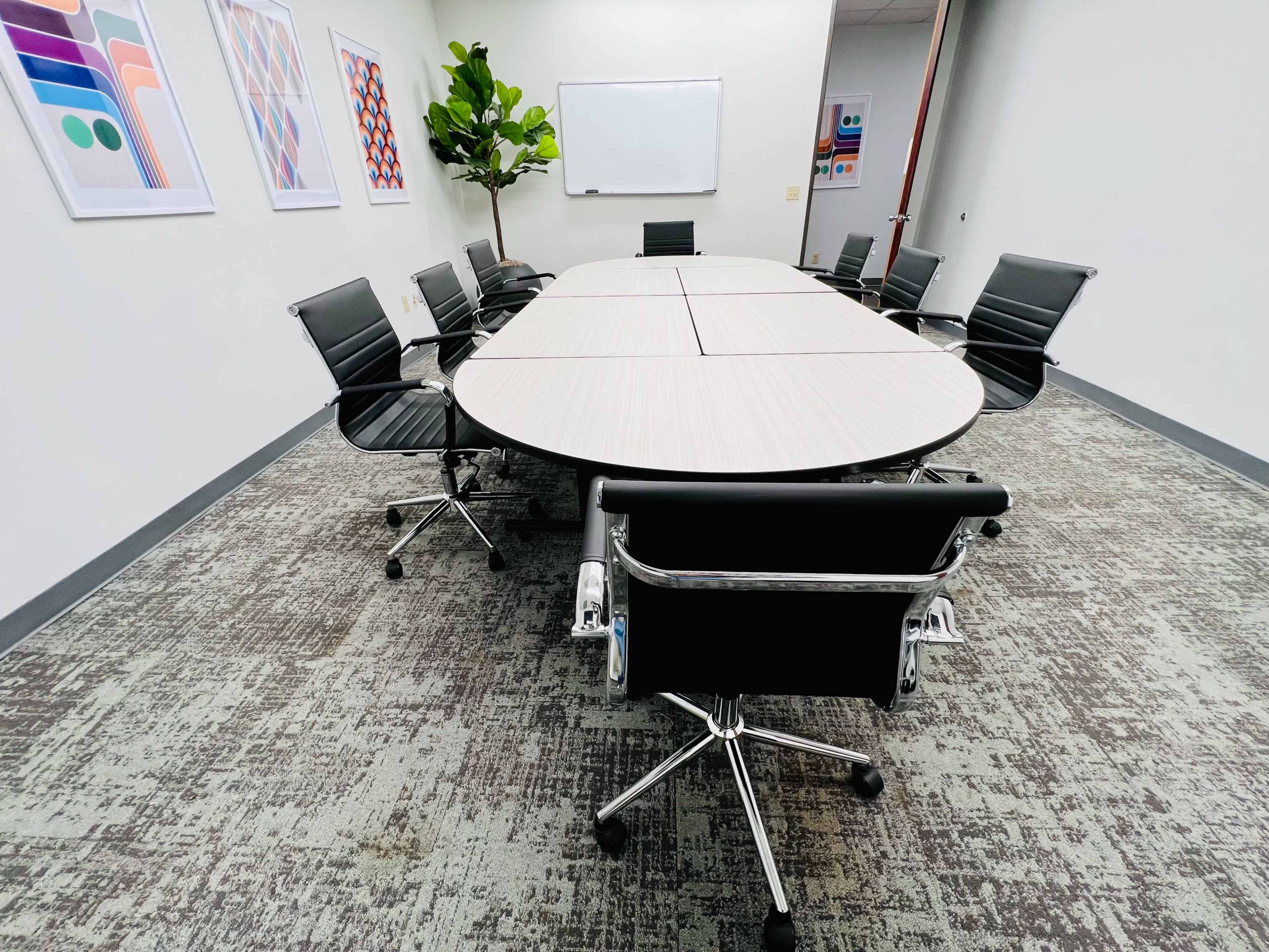 A conference room features a large oval table surrounded by black chairs, with a whiteboard and colorful wall art in the background.