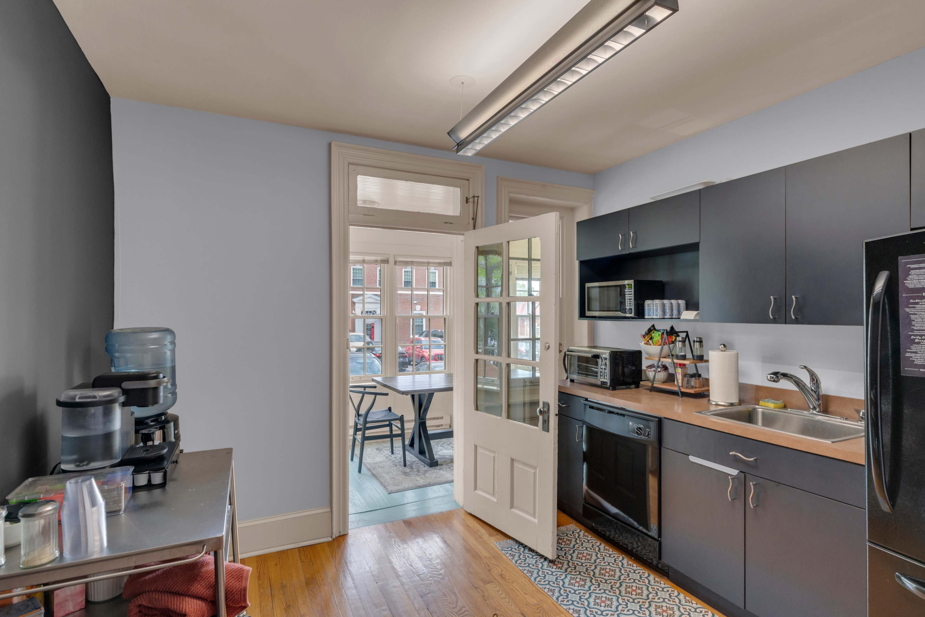 A compact kitchen features dark cabinetry, a countertop with appliances, and a door leading to a dining area with a round table.