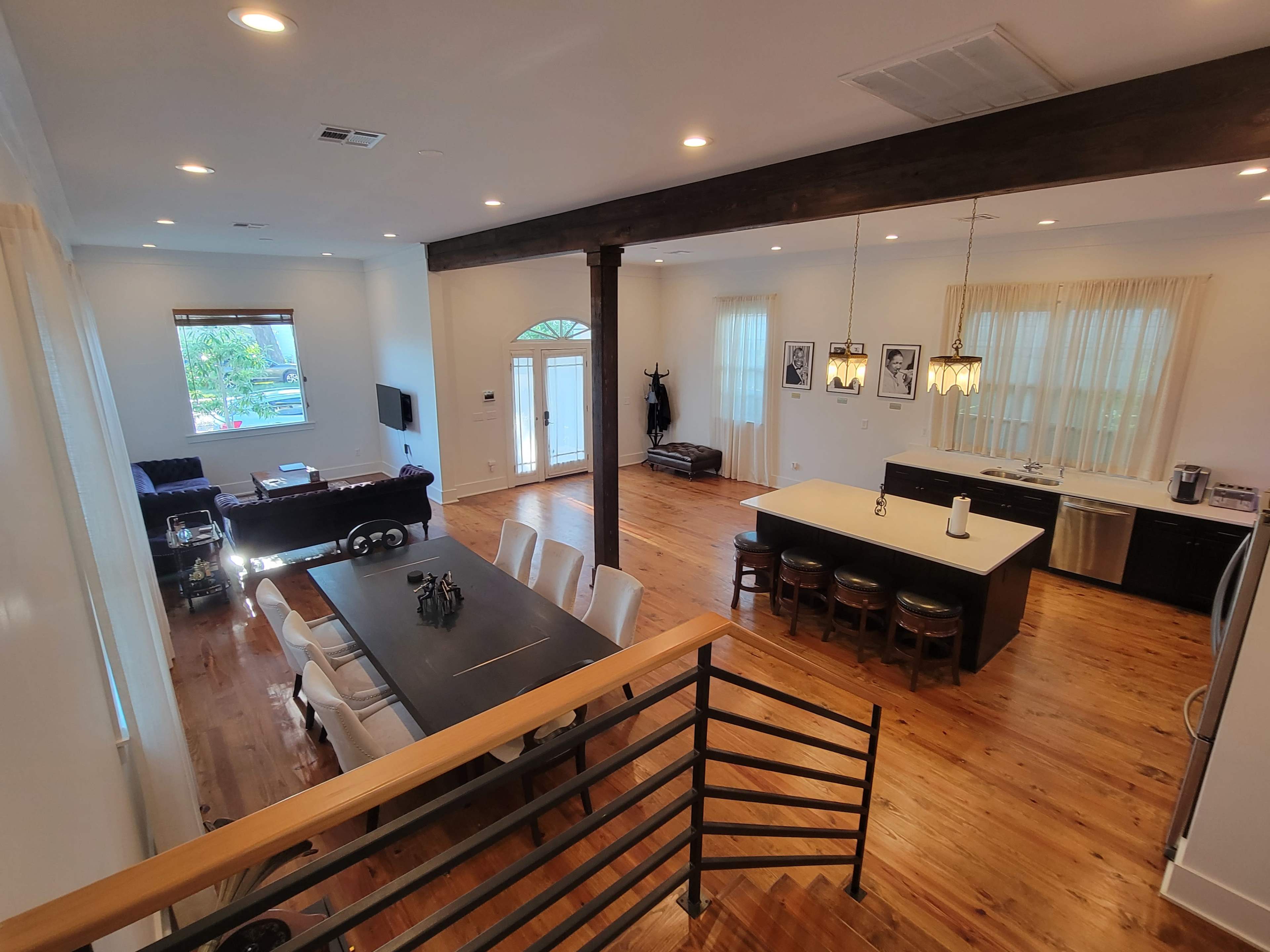 The image shows a bright, open-concept living space featuring a dining area with a long table, a kitchen island with stools, and a cozy seating area with a sofa.