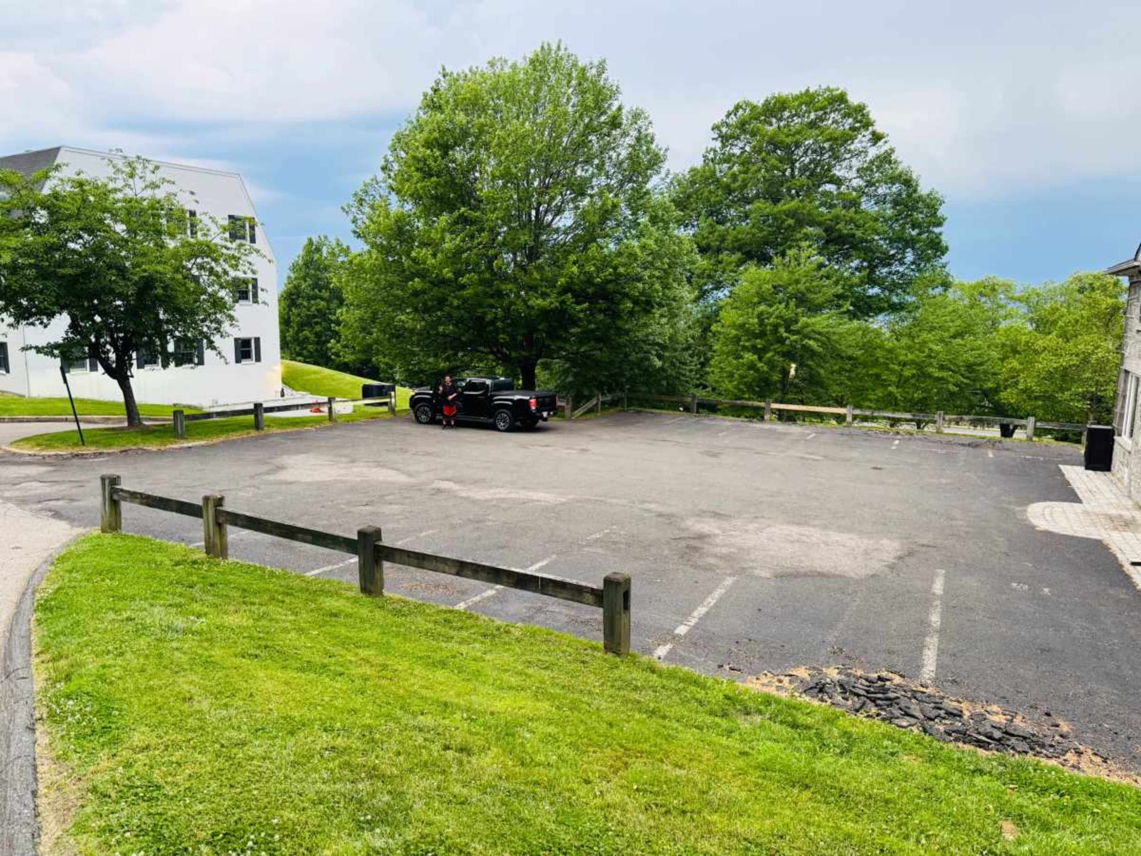 The image shows an empty parking lot surrounded by grass and trees, with a few parked vehicles and a pathway leading to nearby buildings.