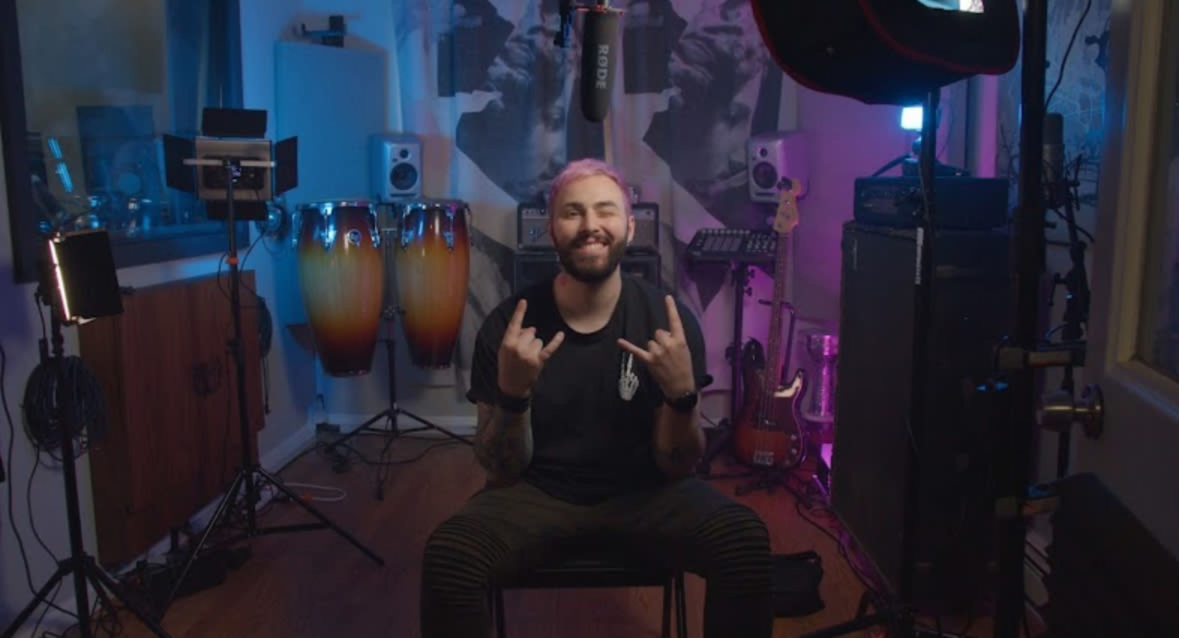 A person with dyed hair is sitting in a recording studio, making a hand gesture while surrounded by musical instruments and studio equipment.