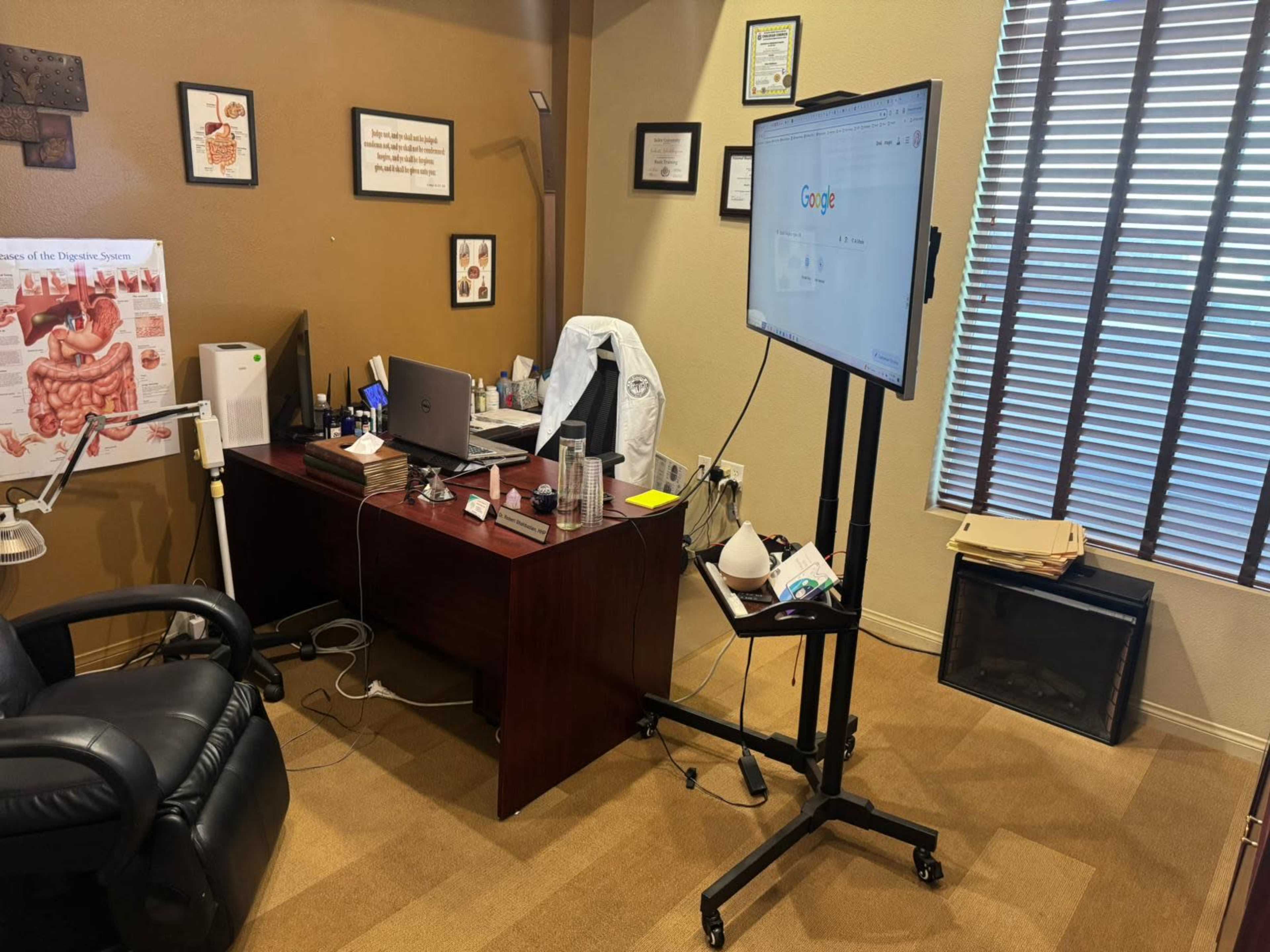The image shows a medical office with a desk, a computer, a large display screen, and a comfortable chair, along with various medical charts and equipment on the walls.