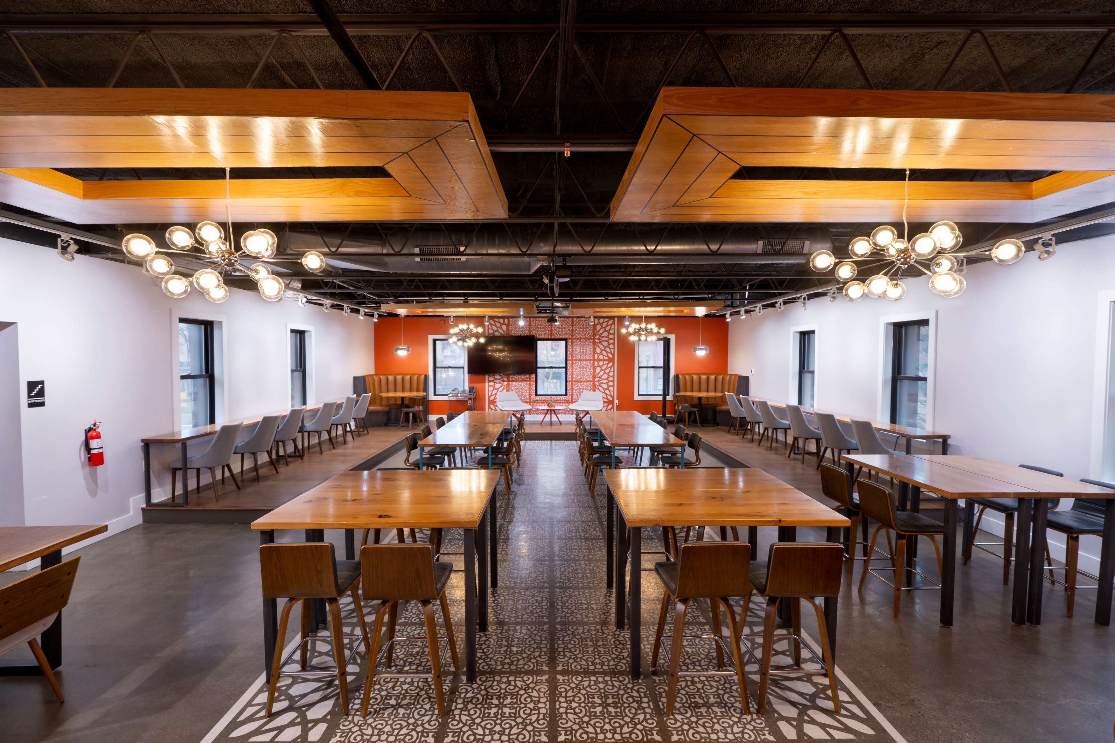 The image shows a spacious dining area with wooden tables and chairs, modern light fixtures, and a decorative wall, arranged for group seating.