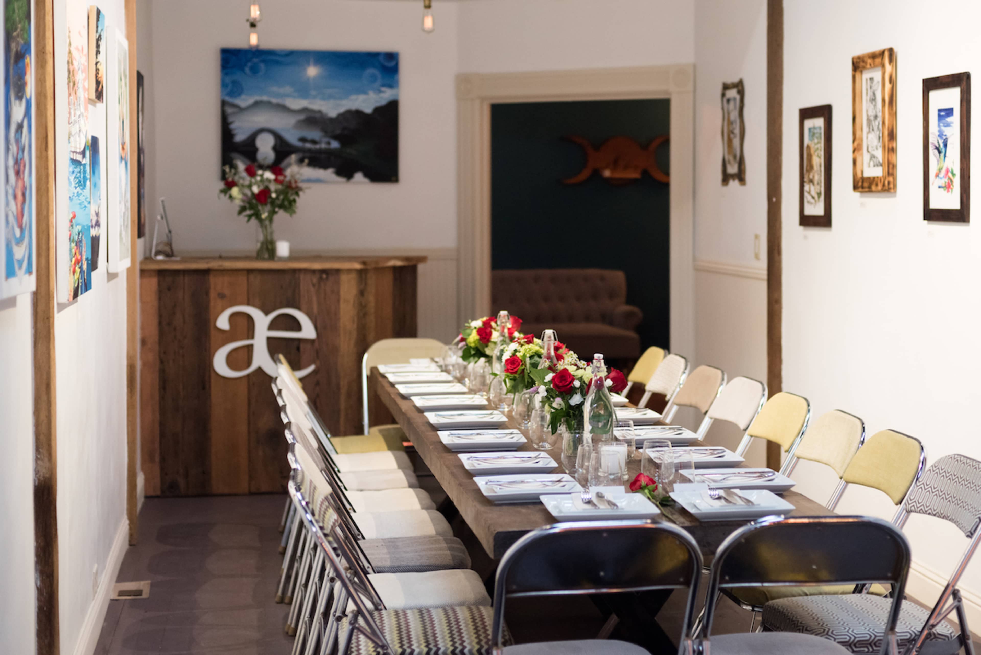 A long wooden table is set with plates and flowers in a brightly lit room featuring artwork on the walls.