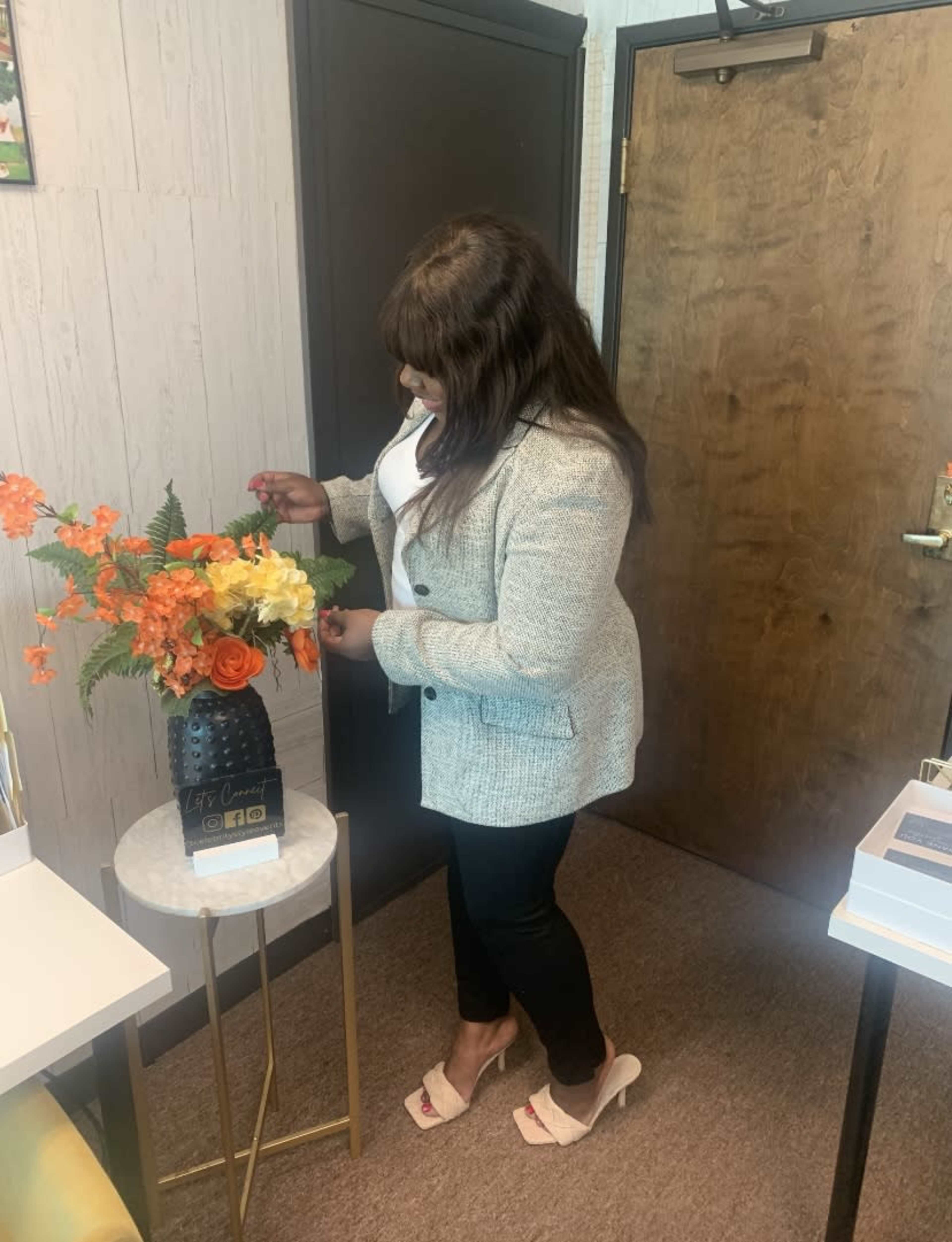 A woman in a light-colored blazer adjusts a floral arrangement on a gold side table near an office door.