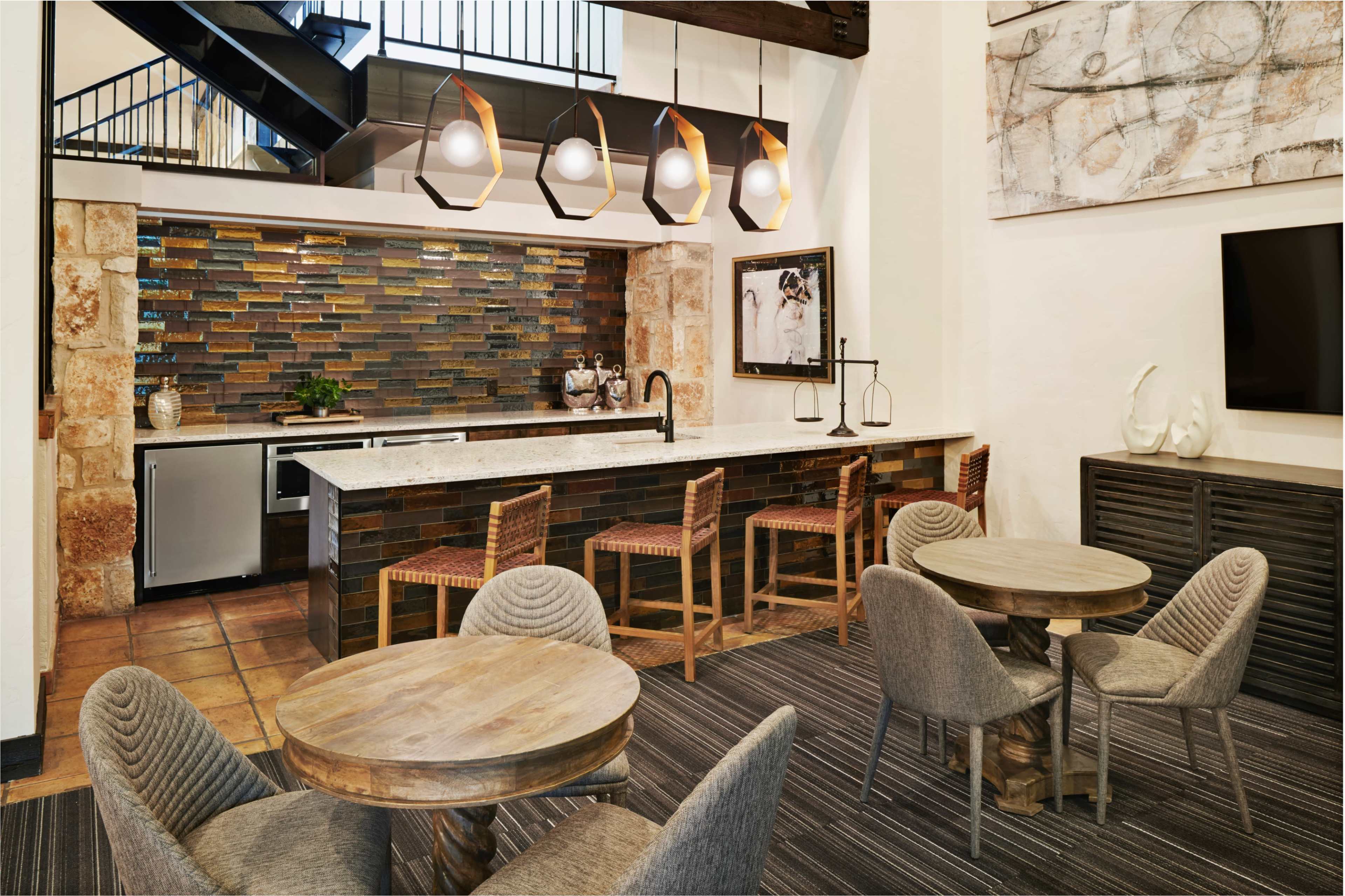 The image shows a modern kitchen area with a granite countertop, bar stools, and a colorful tile backsplash, surrounded by dining tables and decorative lighting fixtures.