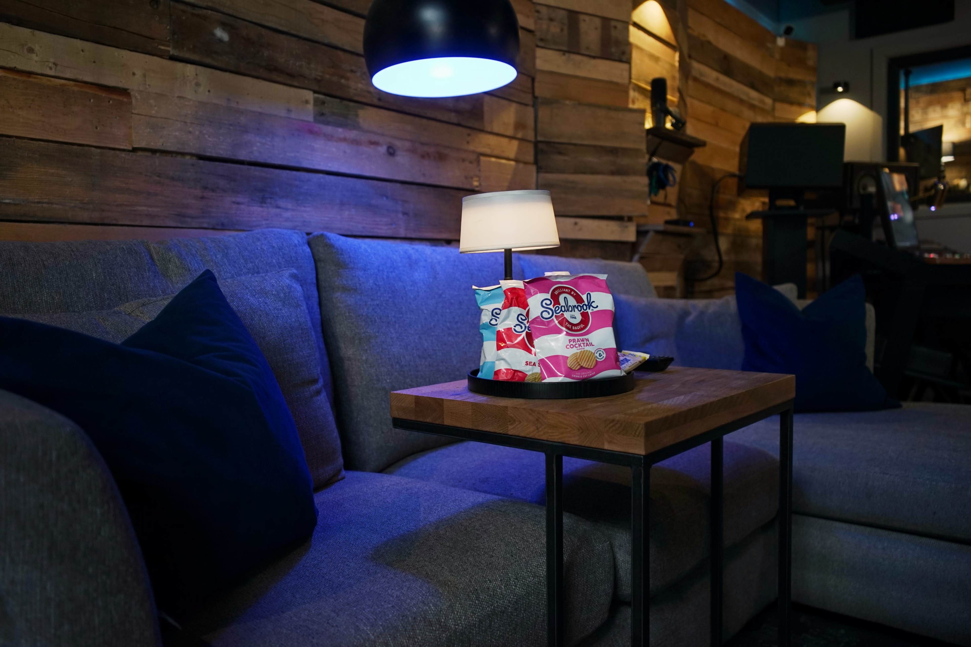 A cozy living room scene features a couch with navy blue pillows, a wooden table holding snacks, and a lamp under a hanging light fixture against a backdrop of wooden wall panels.