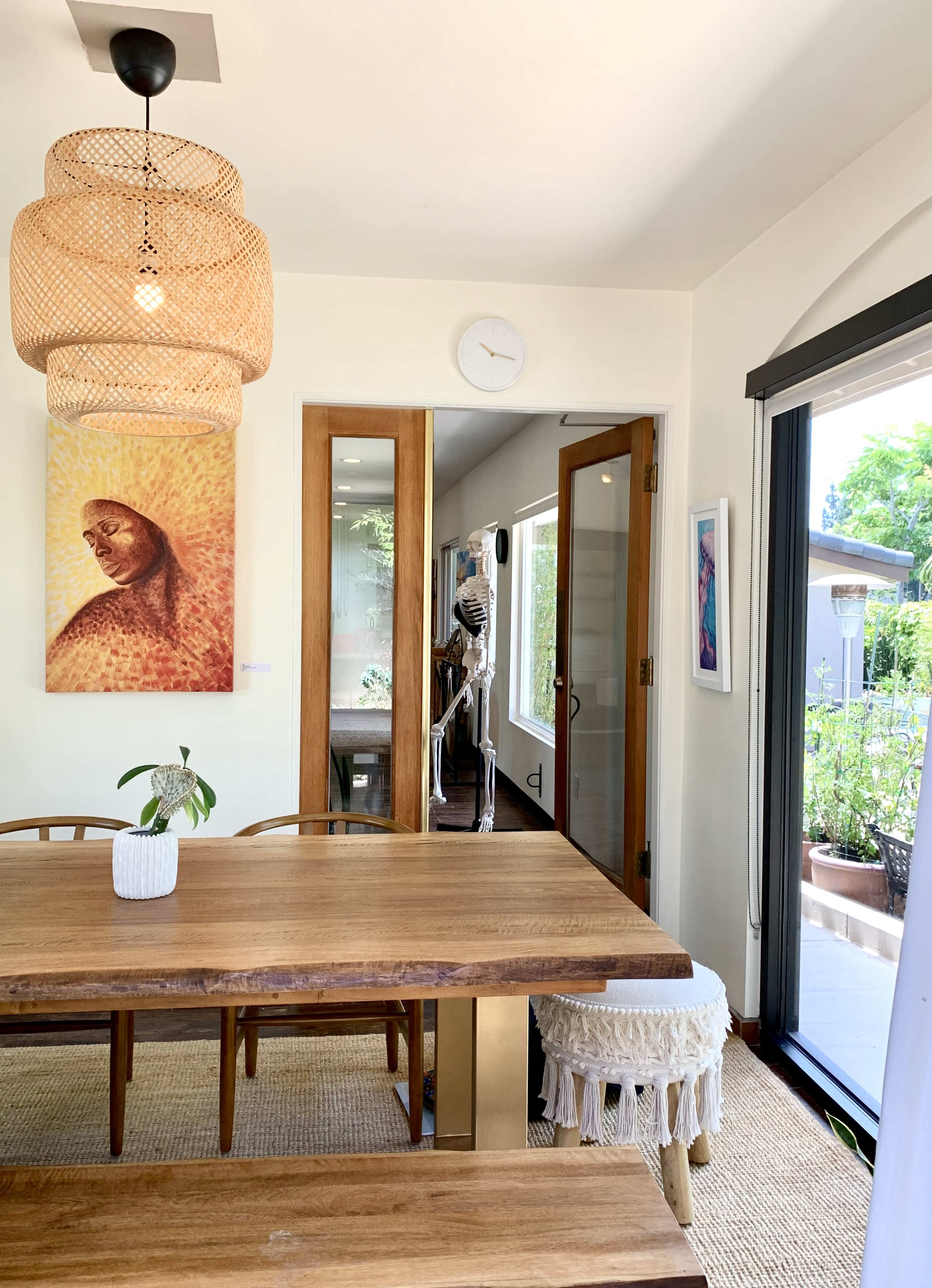 A dining area with a wooden table, a woven pendant light, and a doorway leading to another room where a skeleton figure is partially visible.
