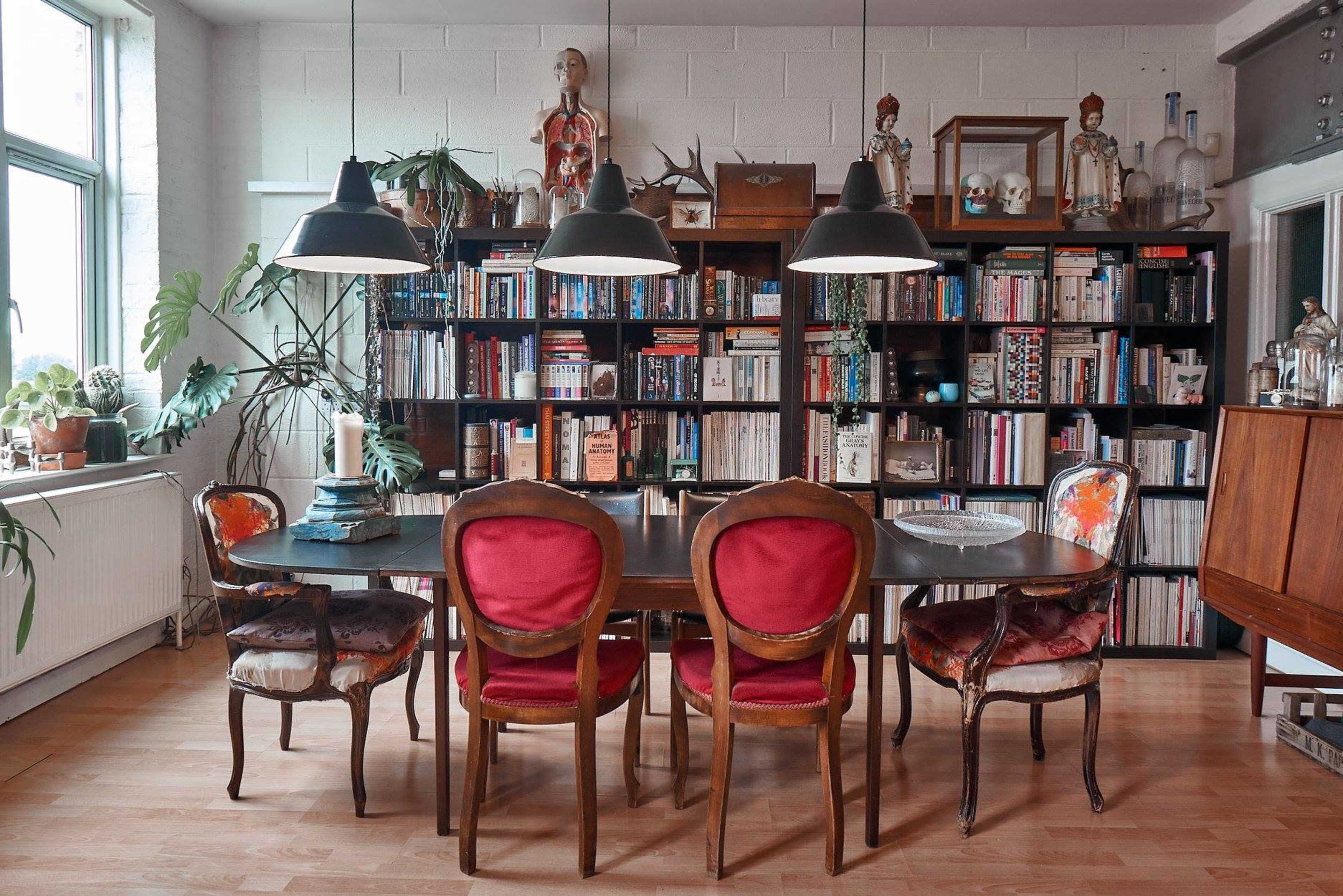 A dining room features a large table surrounded by six chairs, with a bookshelf filled with books and decorative items in the background.