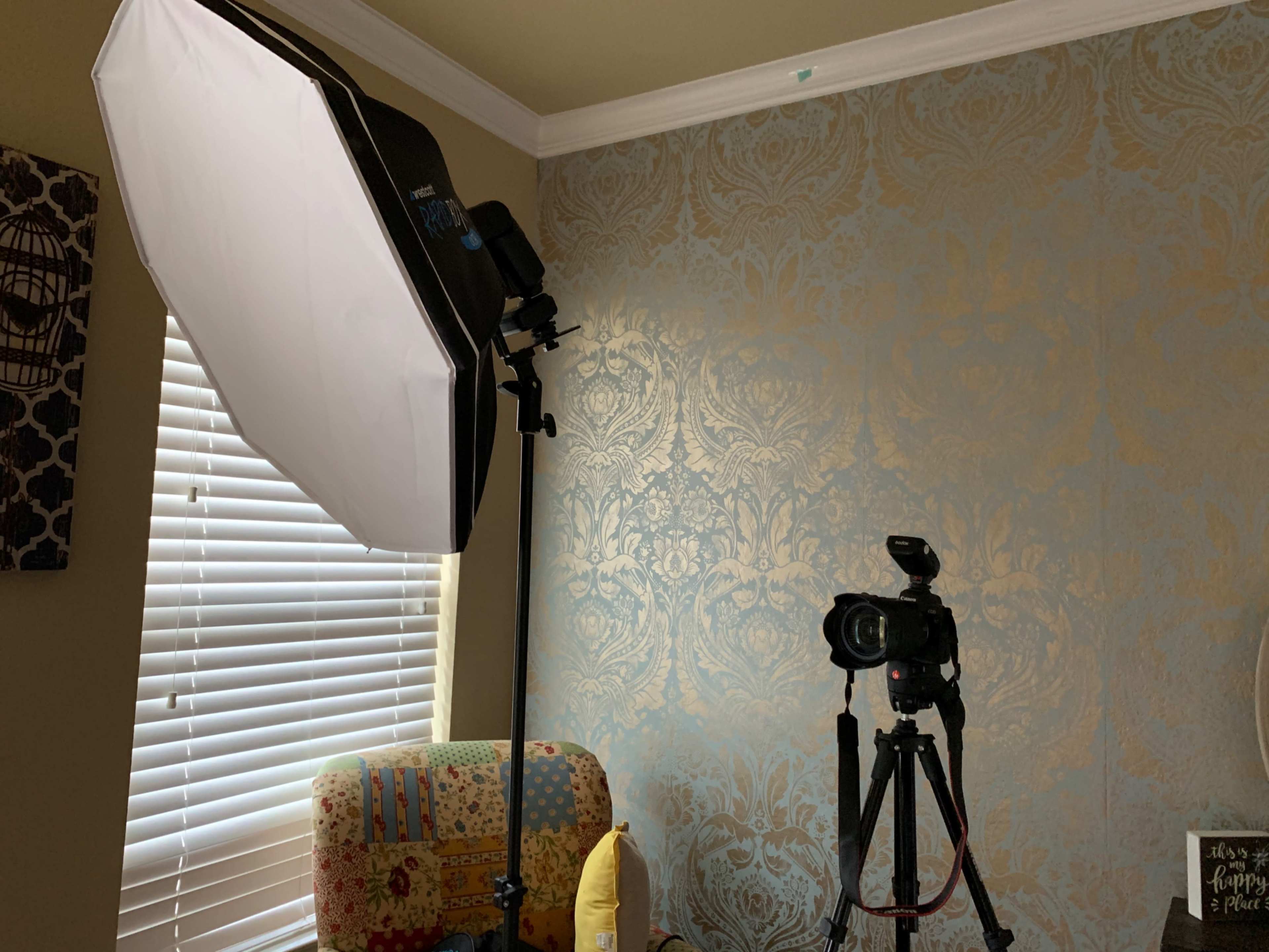 A photography setup features a softbox light and a camera mounted on a tripod, placed near patterned wallpaper and a colorful chair.
