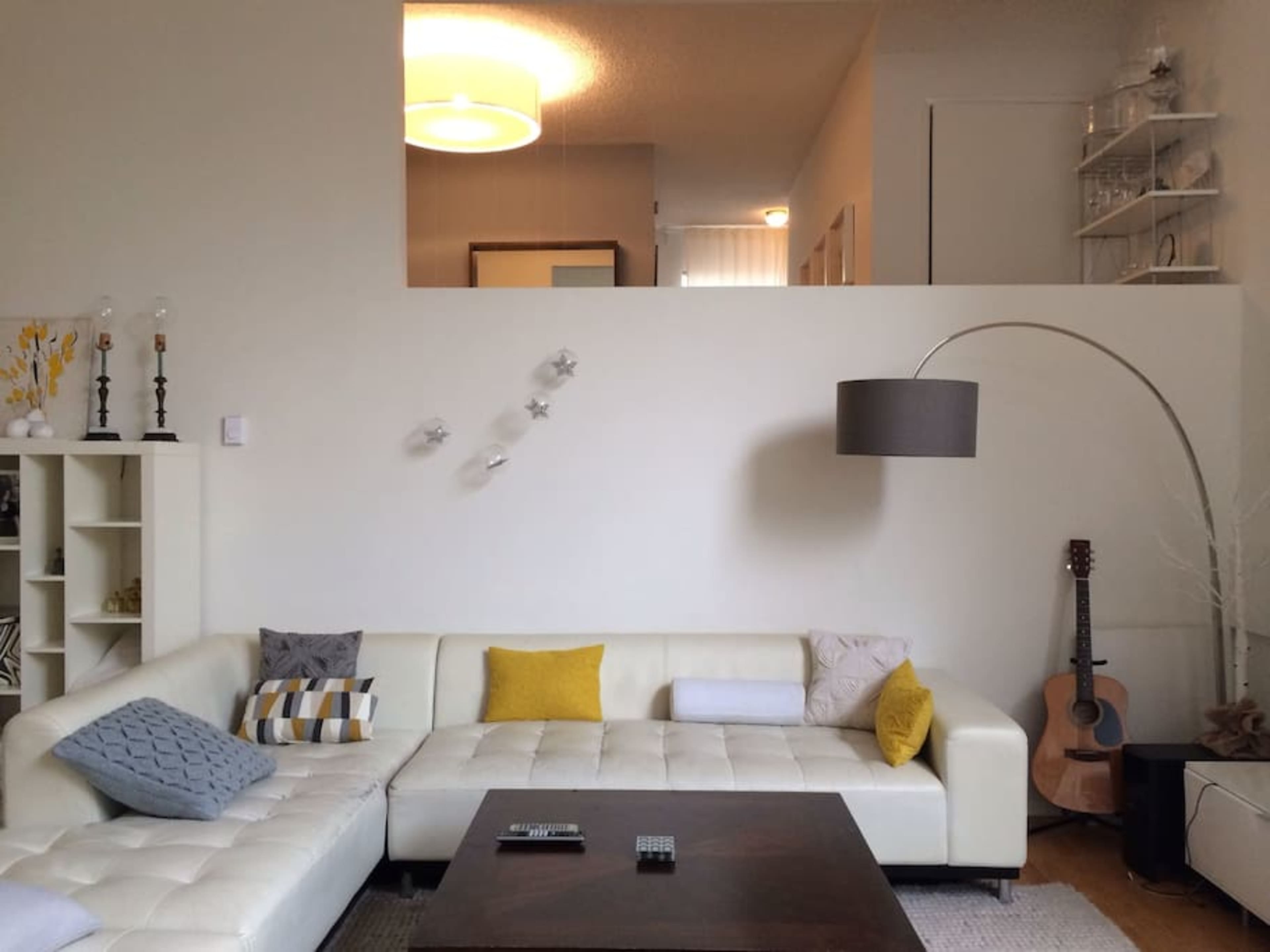 A modern living room features a white sectional sofa with colorful cushions, a wooden coffee table, and a guitar in the corner, with a staircase visible in the background.