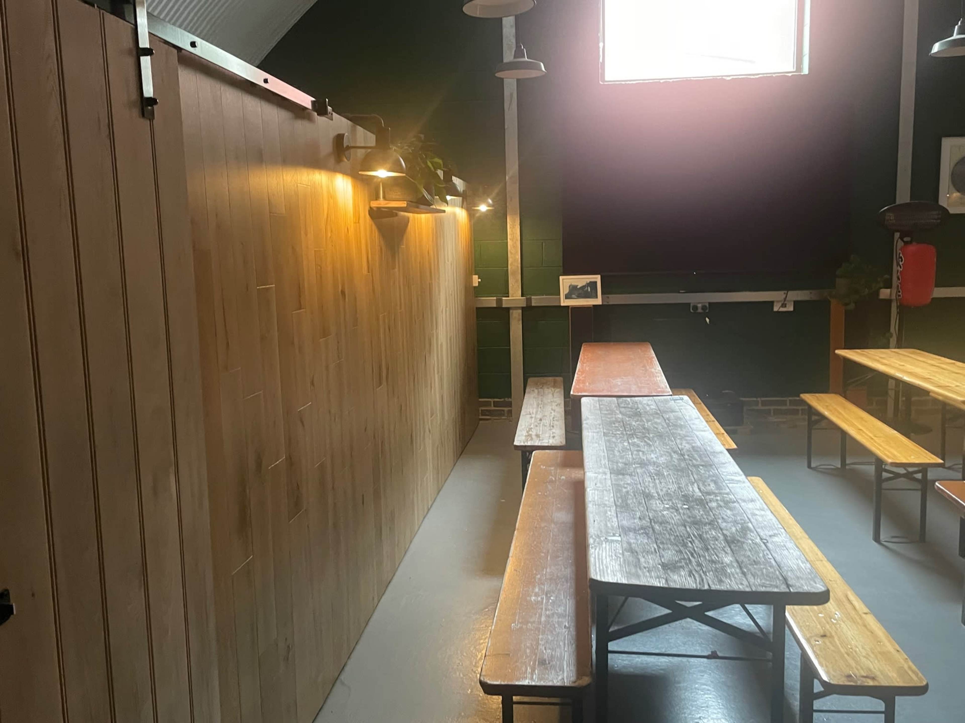 The image shows a sparsely furnished room with wooden benches and tables, illuminated by warm light from a wall-mounted fixture.