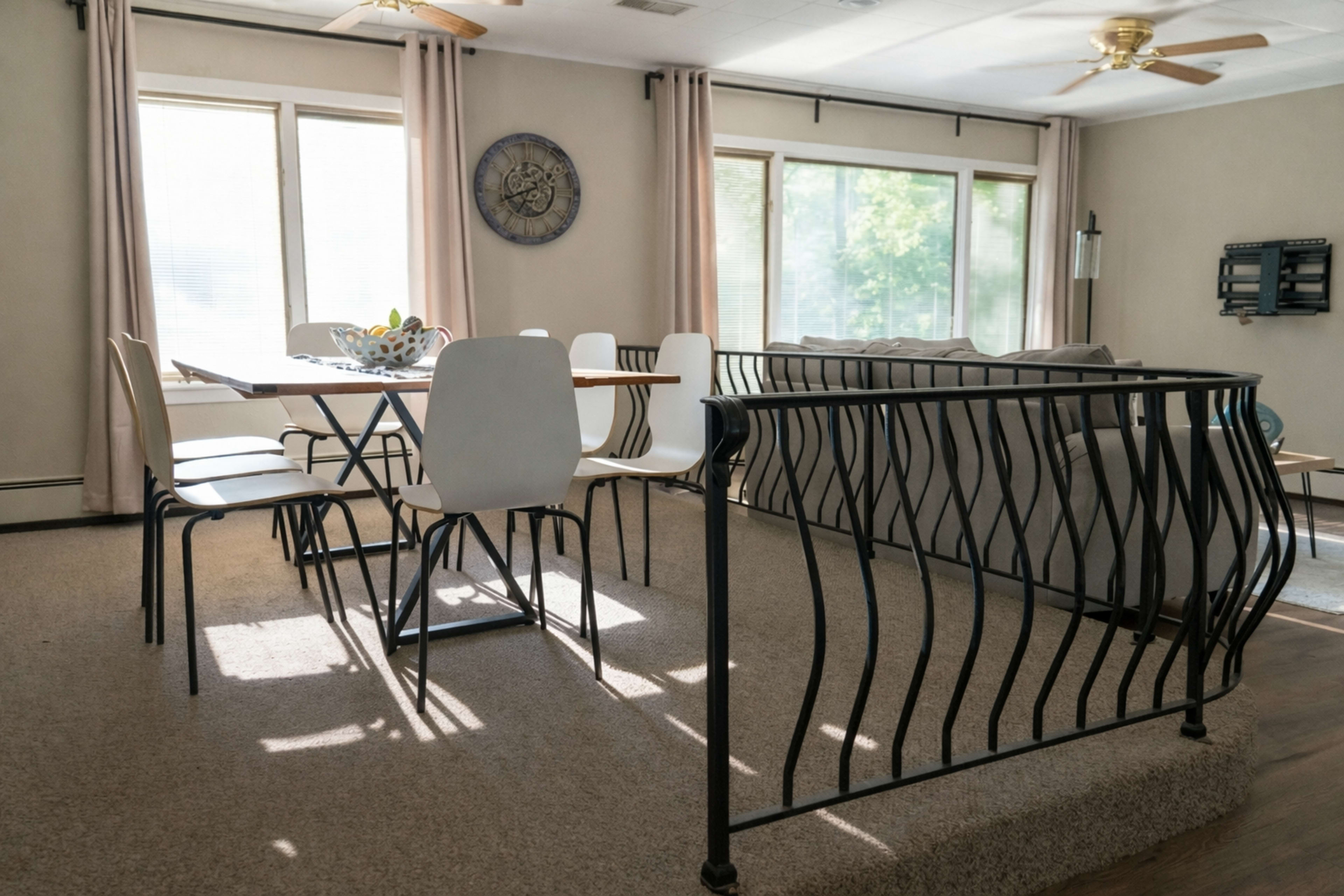 The image shows a living room with a dining area featuring a table and chairs, large windows allowing natural light, and a railing separating the two spaces.