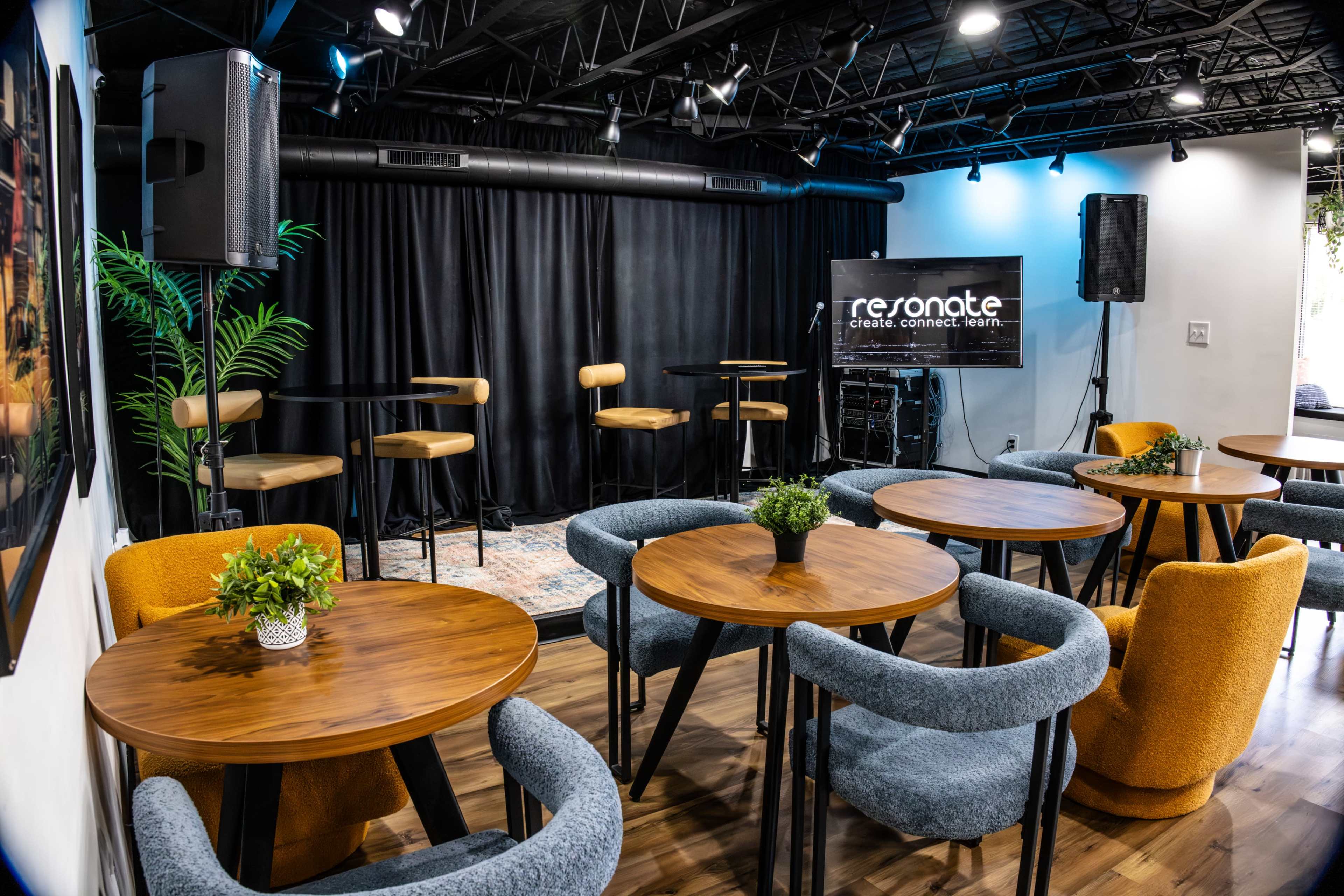 The image shows a small, modern event space with wooden tables and chairs arranged in front of a stage featuring tall speakers and bar stools.
