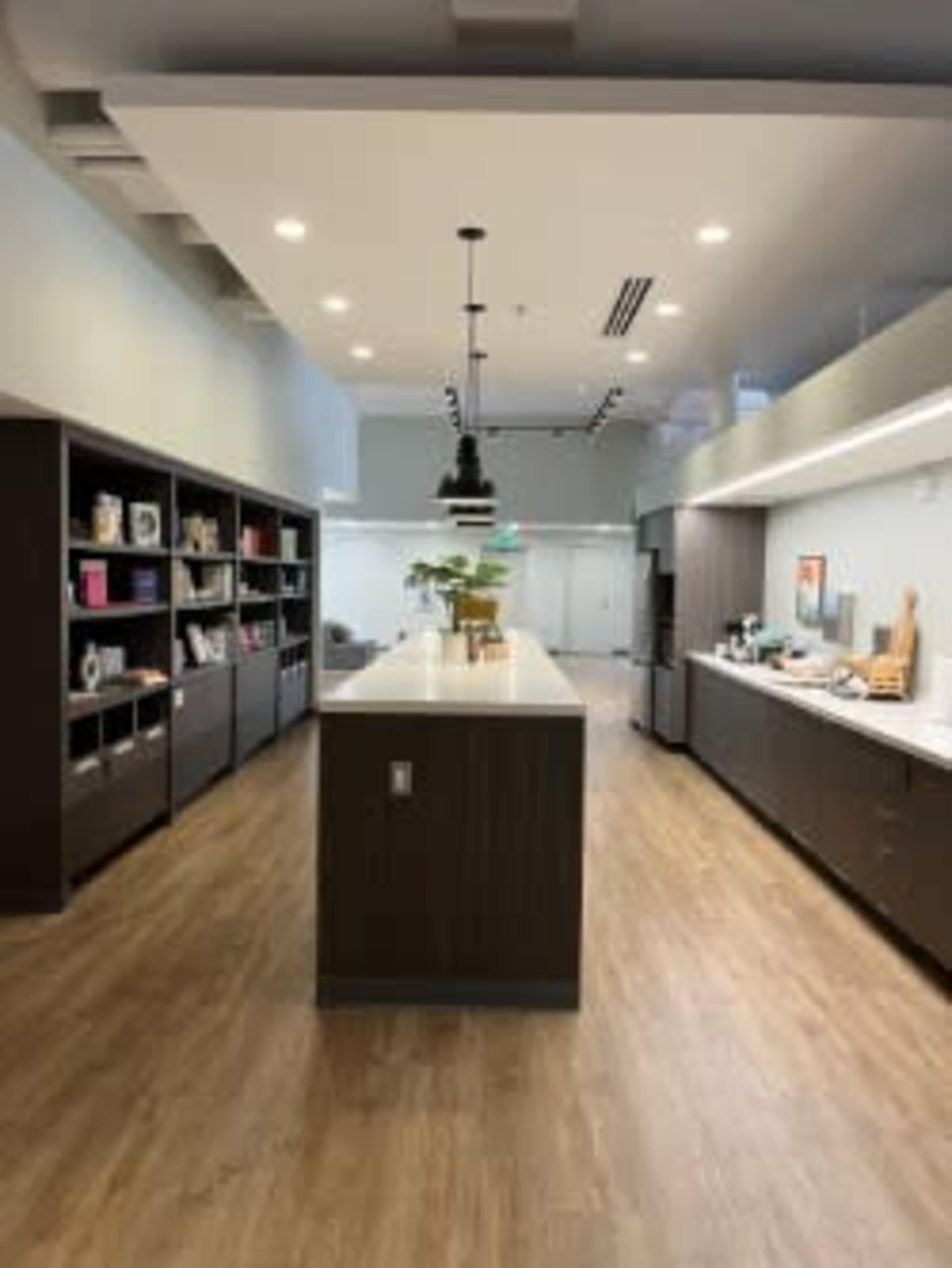 A modern office kitchen area features a central island with a countertop, shelving displaying books and decorative items, and a range of appliances along one side.