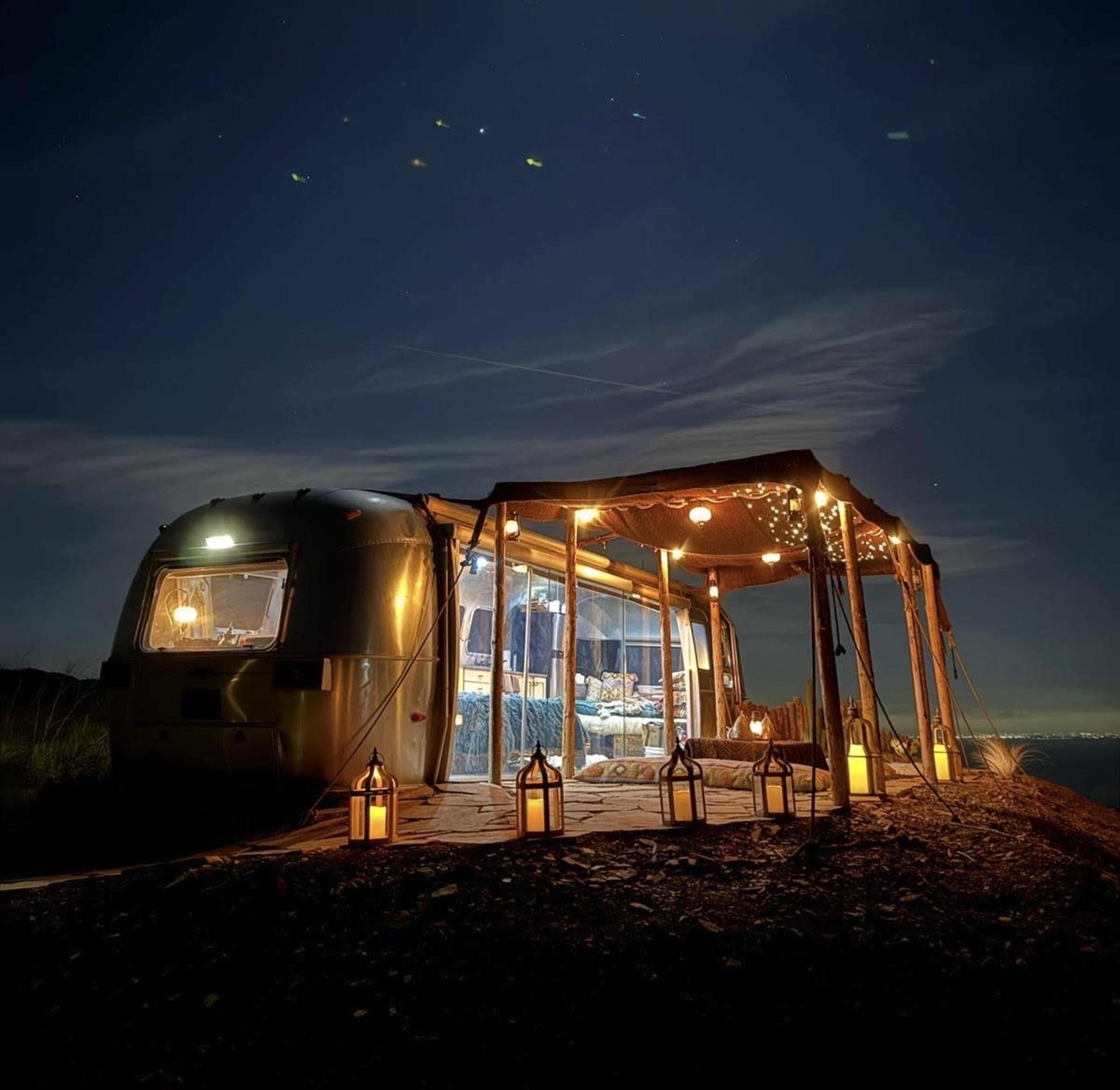 A retro Airstream trailer is illuminated at night under a canopy adorned with string lights and lanterns, set against a dark sky.