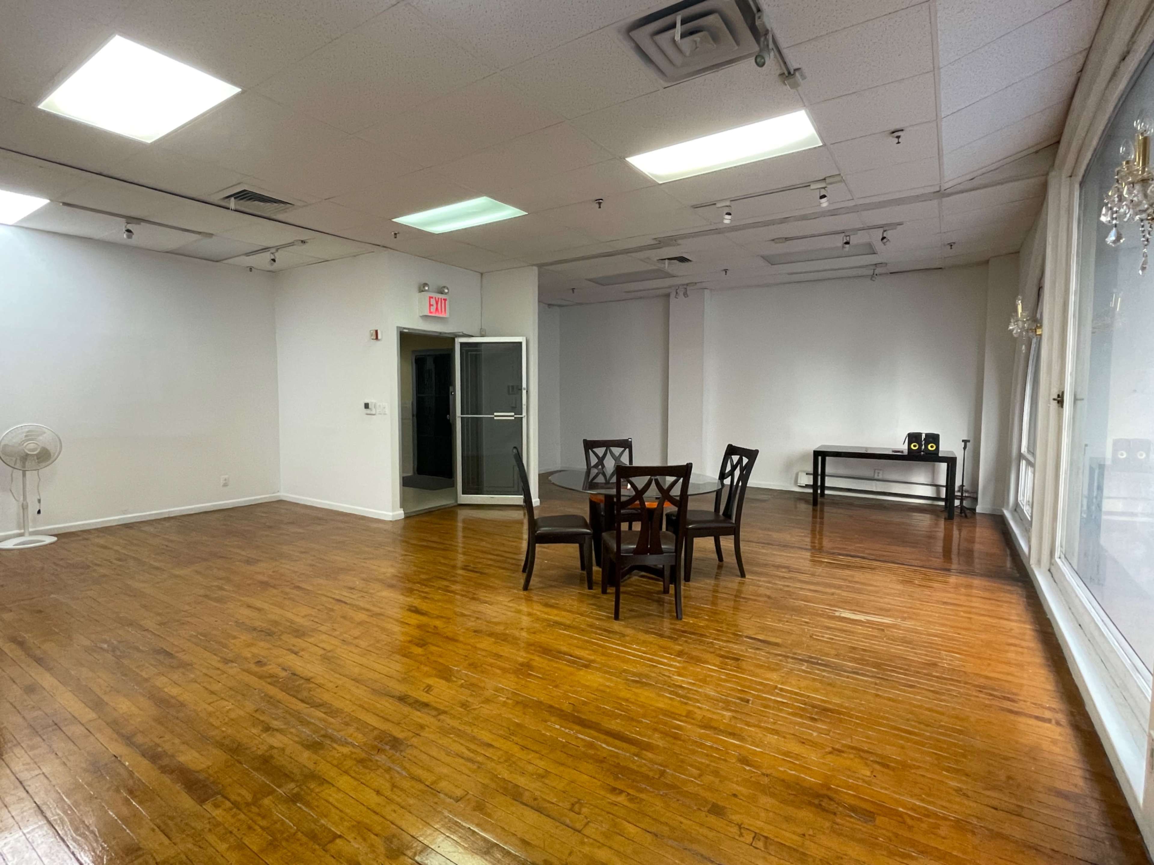 The image shows a spacious, empty room with polished wooden floors, a round table with chairs in the center, and a side table with speakers against a wall.