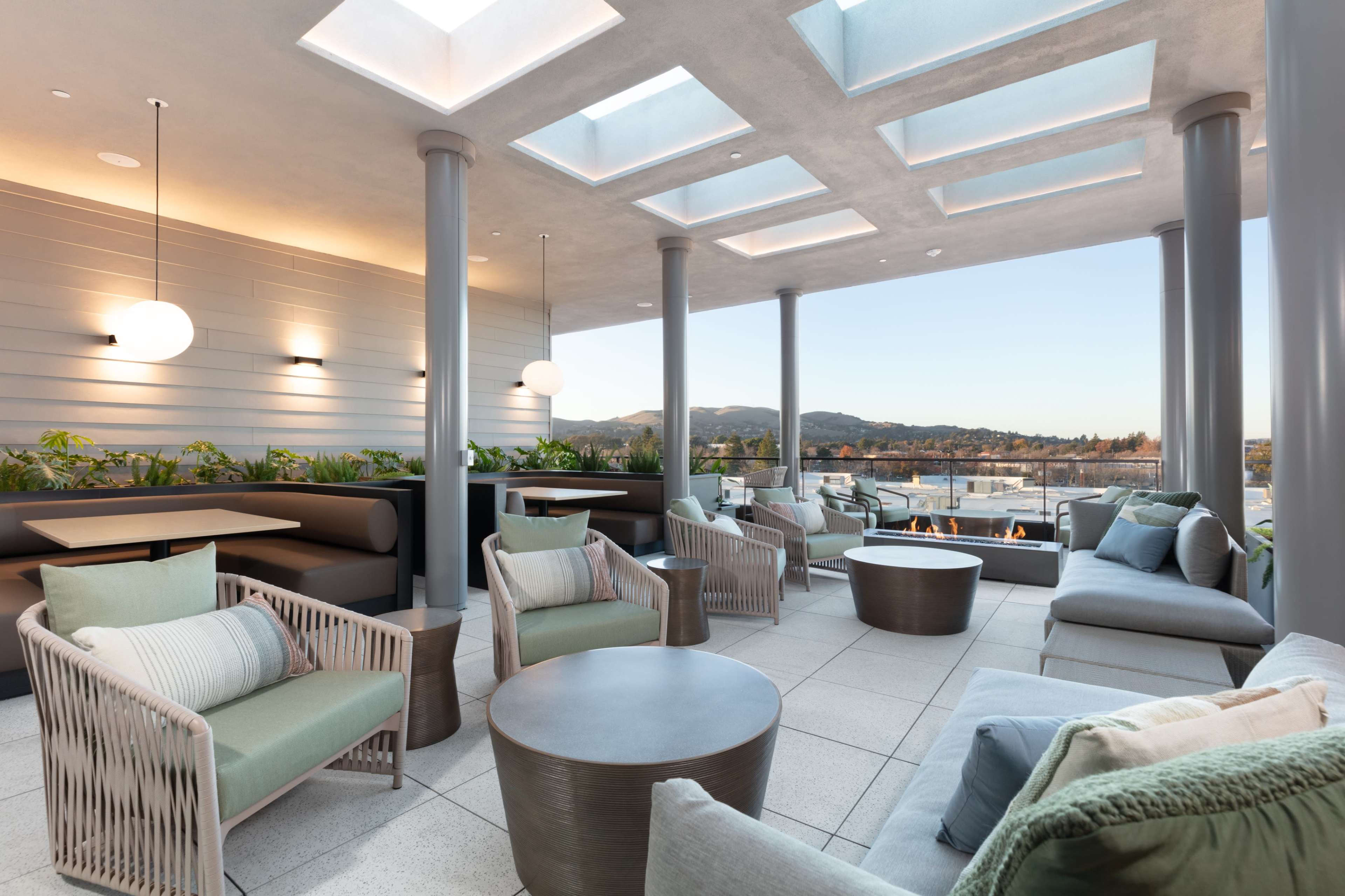 The image shows a modern rooftop lounge area with comfortable seating, a fire pit, and glass ceiling panels allowing natural light to enter.