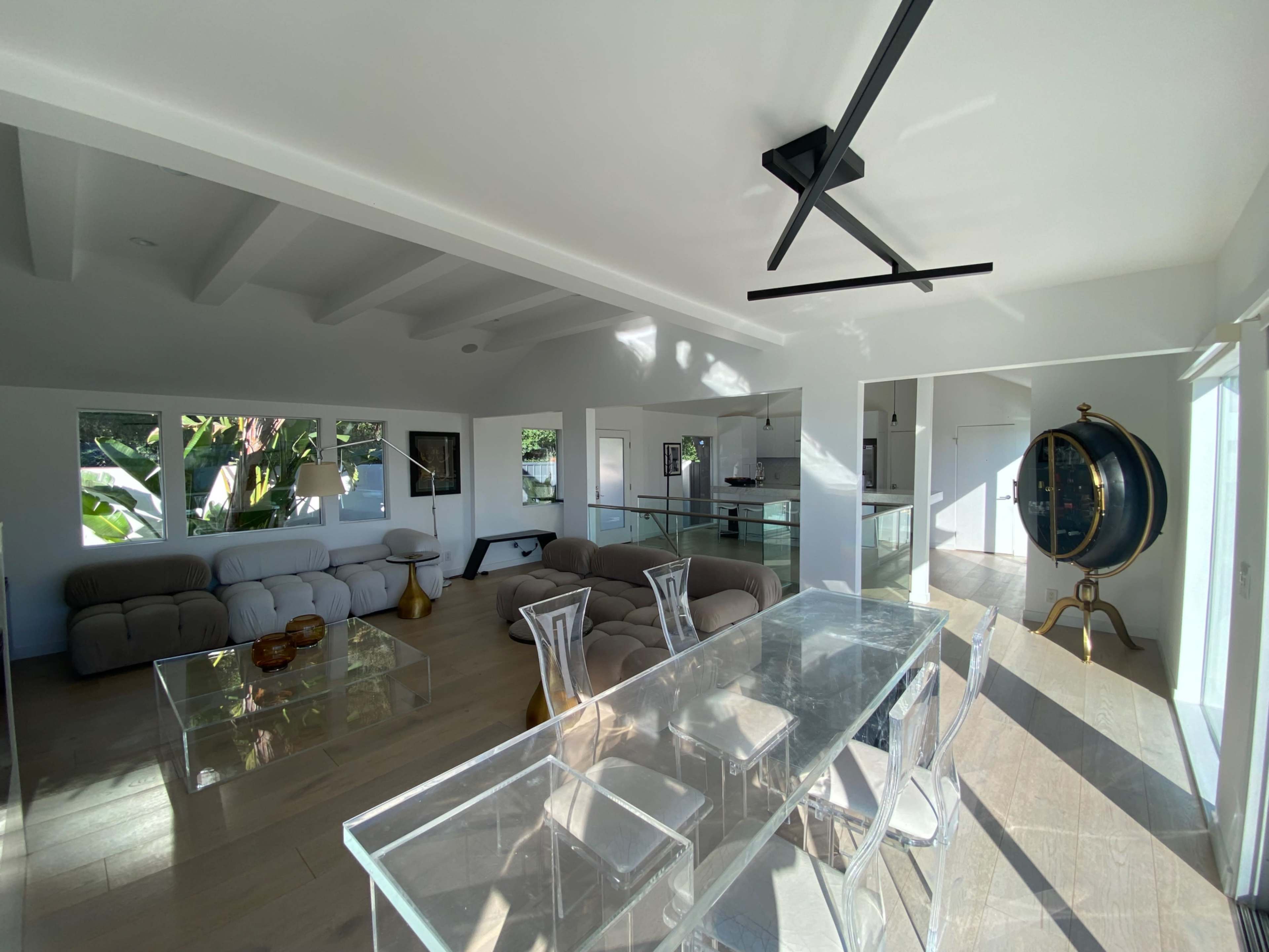 The image shows a modern open-concept living area featuring a light-colored sofa, a glass dining table, and large windows providing natural light.
