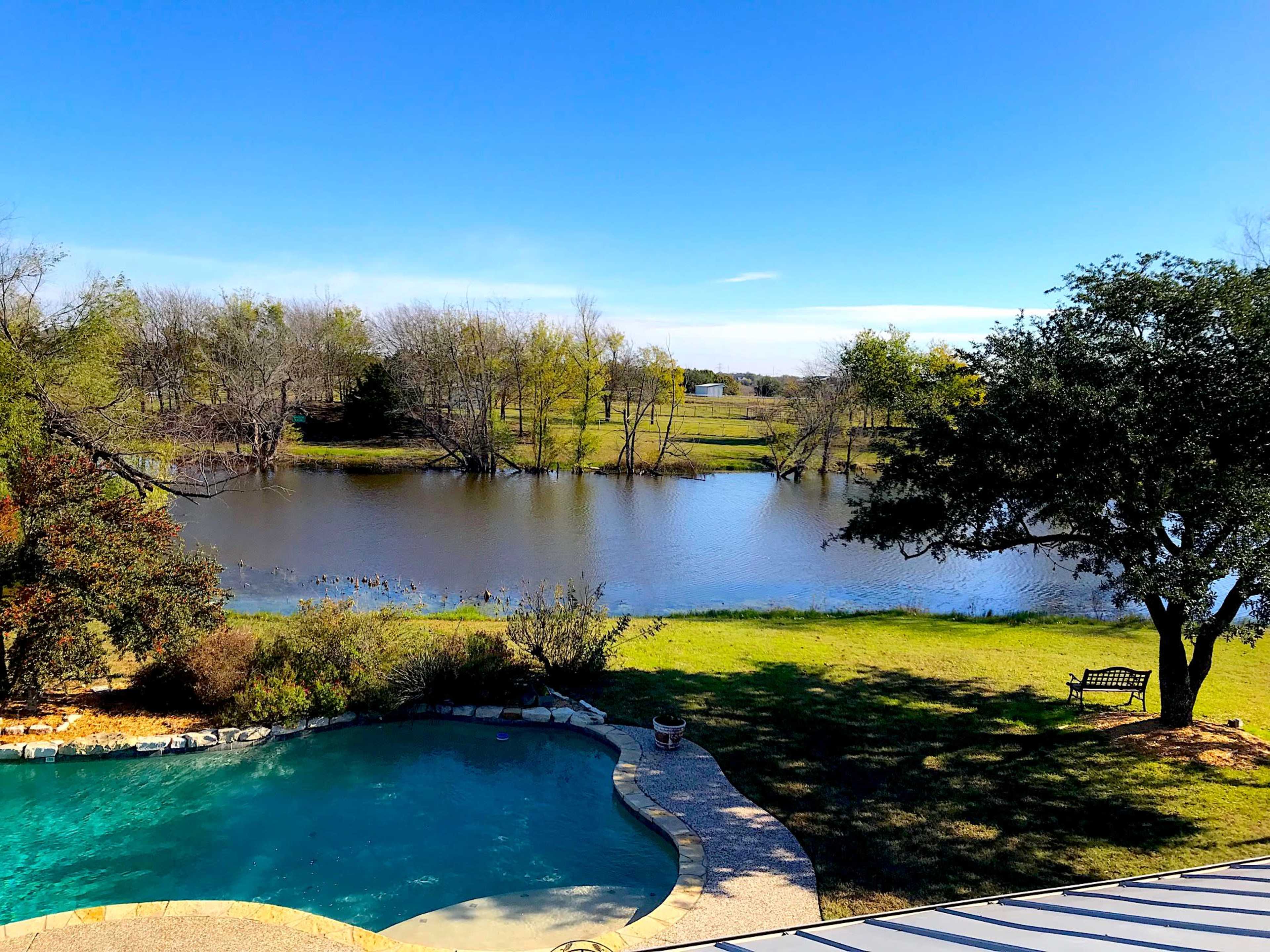 A backyard view features a swimming pool adjacent to a calm pond surrounded by trees and grassy areas.