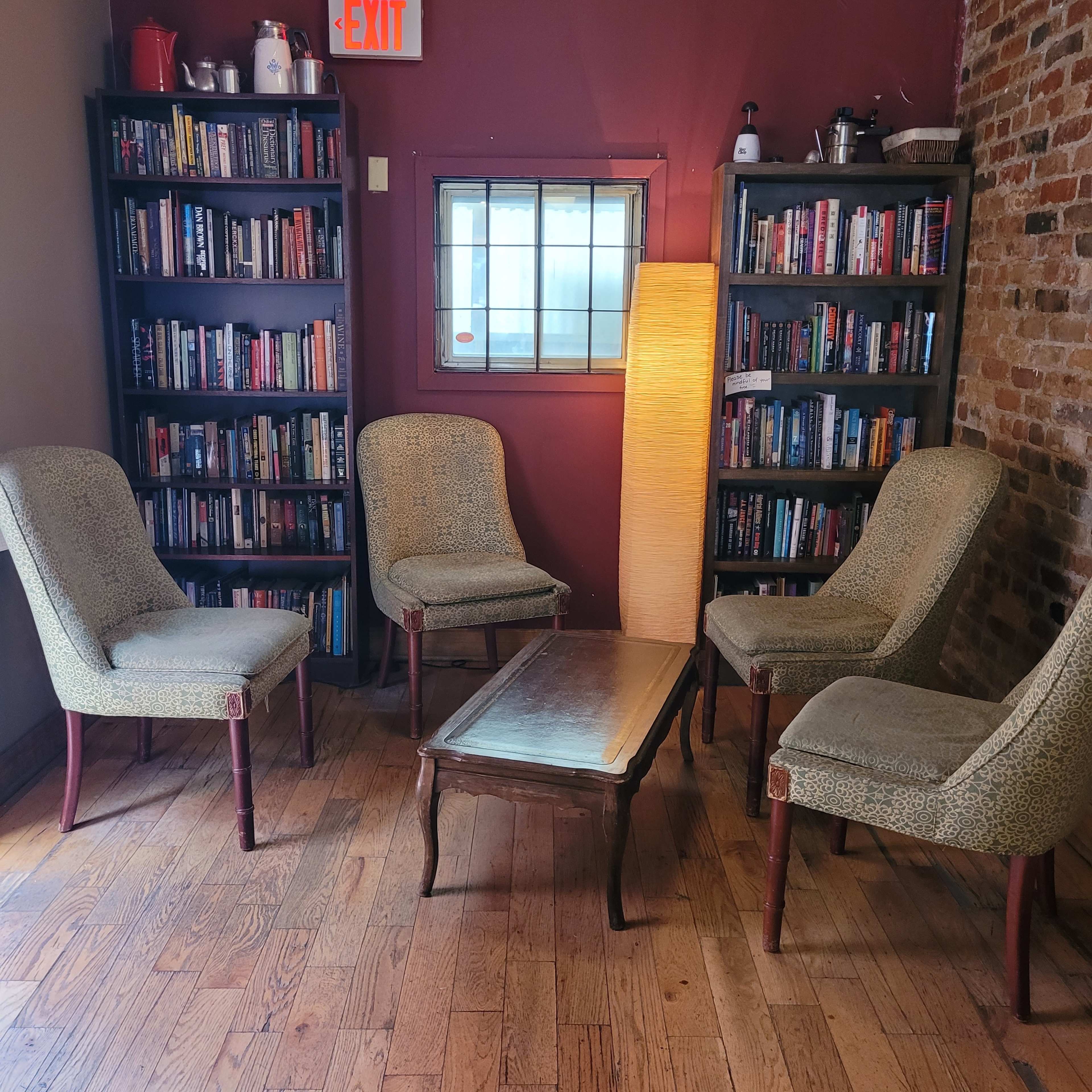 A cozy reading corner with four upholstered chairs arranged around a wooden coffee table, flanked by shelves filled with books, and a floor lamp providing soft lighting.