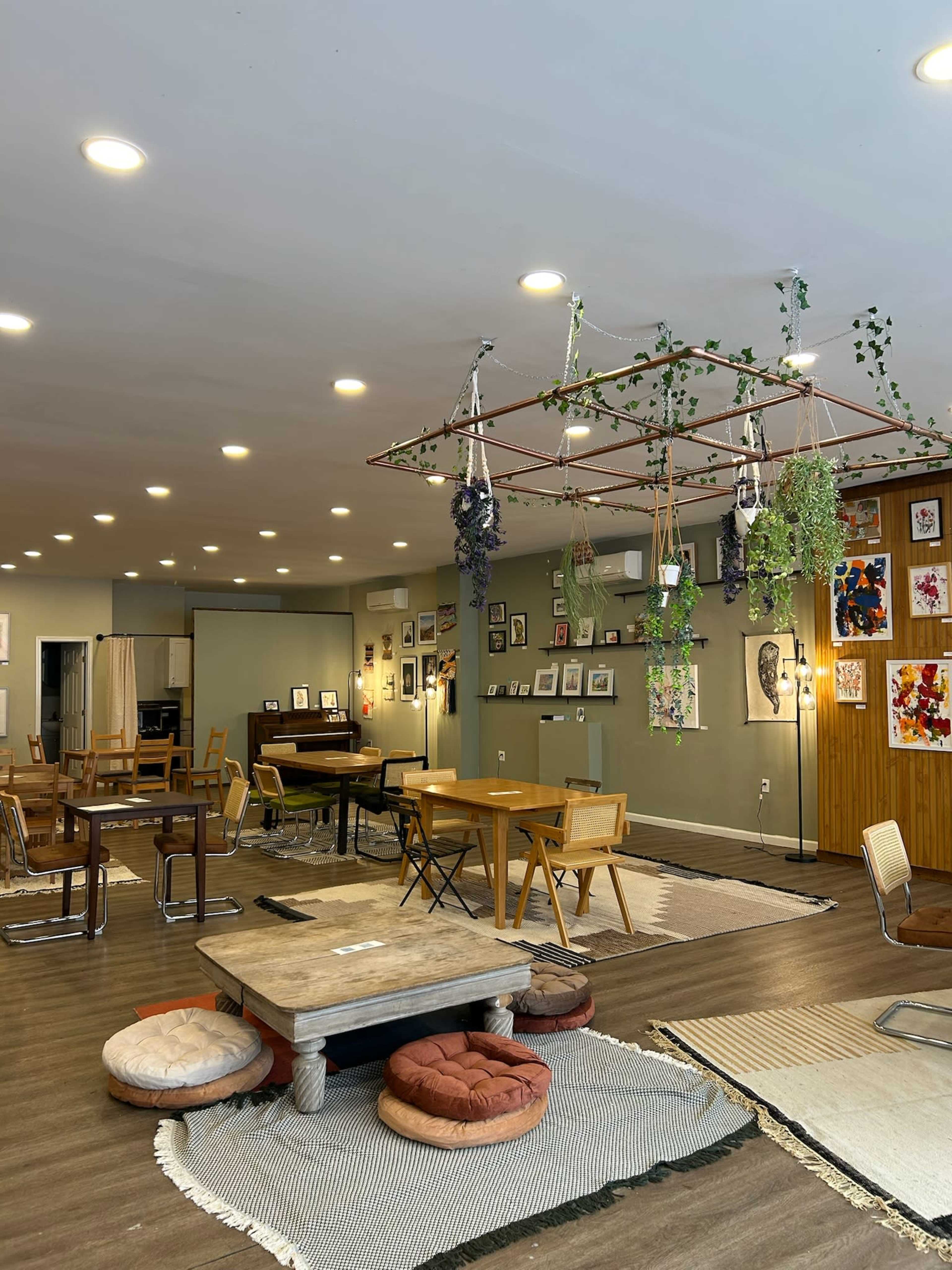 A spacious café interior with various seating arrangements, including wooden tables, cushions on the floor, and hanging plants from the ceiling.