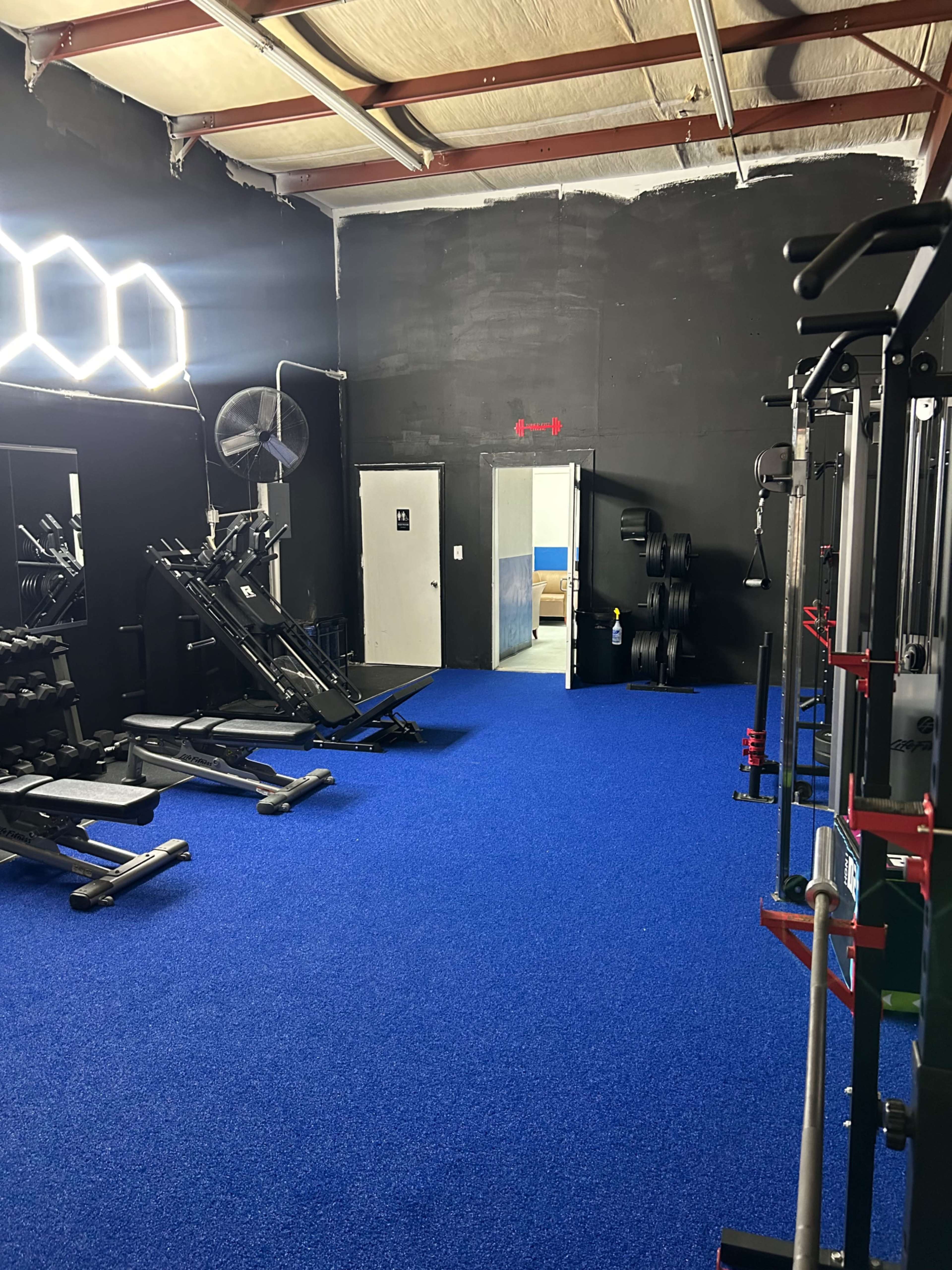 The image shows a gym interior featuring exercise equipment, blue carpet flooring, and an open door leading to another room.