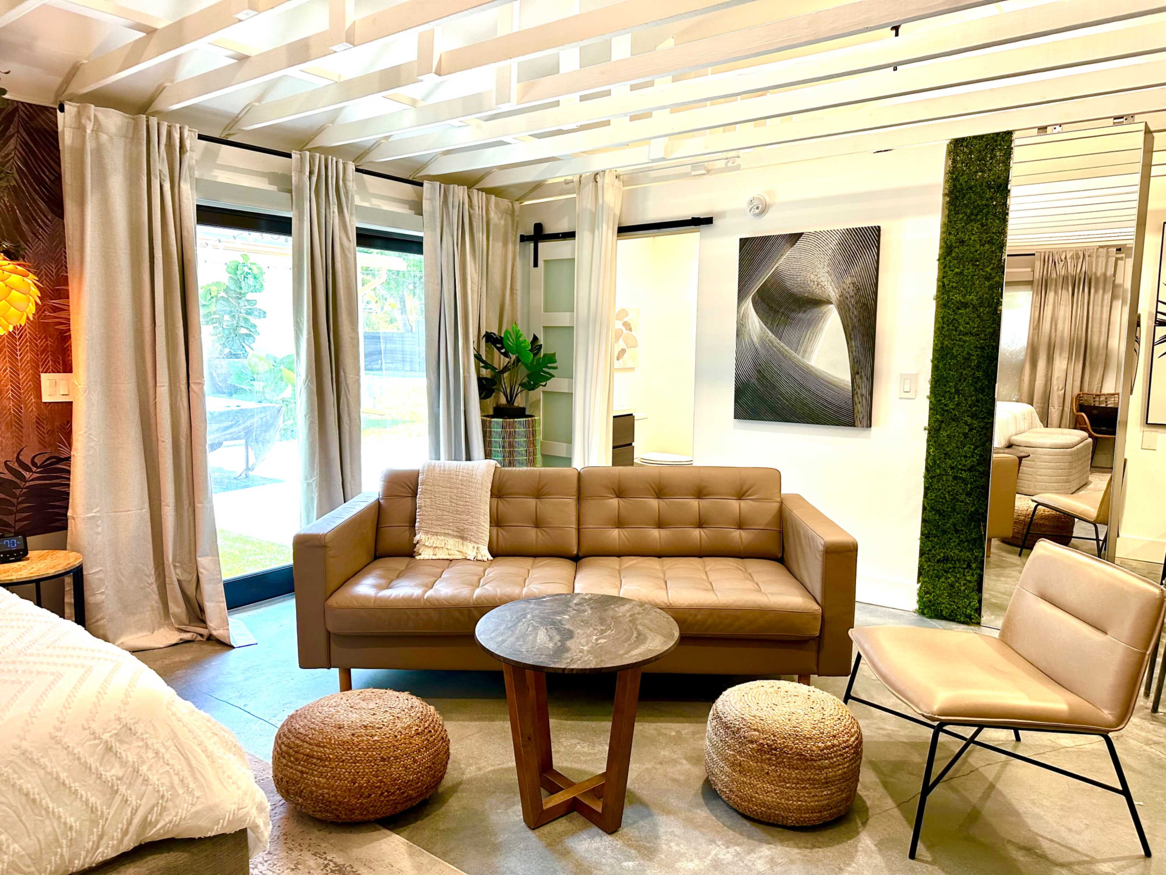 A modern interior living space featuring a brown leather sofa, a round coffee table, woven poufs, and large windows with sheer curtains.