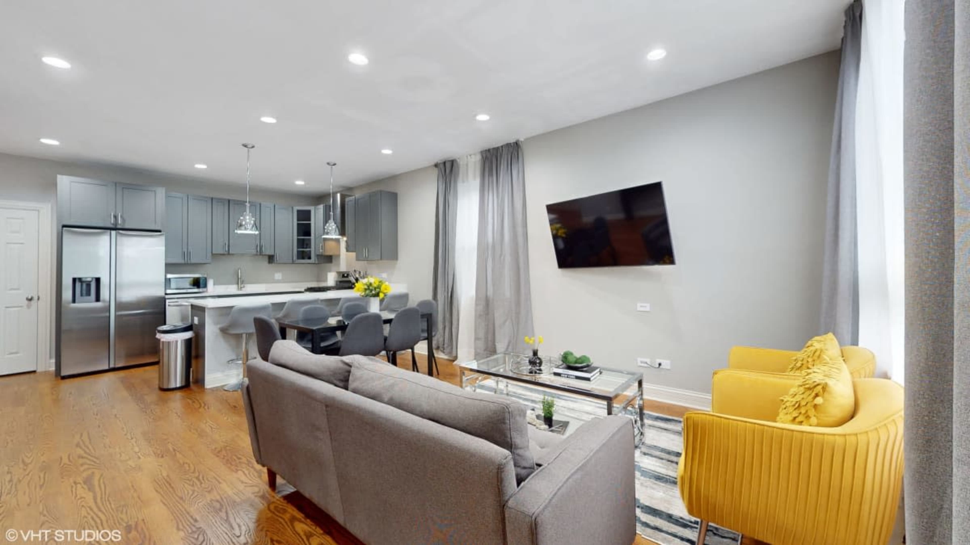 The image shows a modern living area with a gray sofa, yellow accent chairs, a glass coffee table, and a kitchen area featuring stainless steel appliances and a large window.