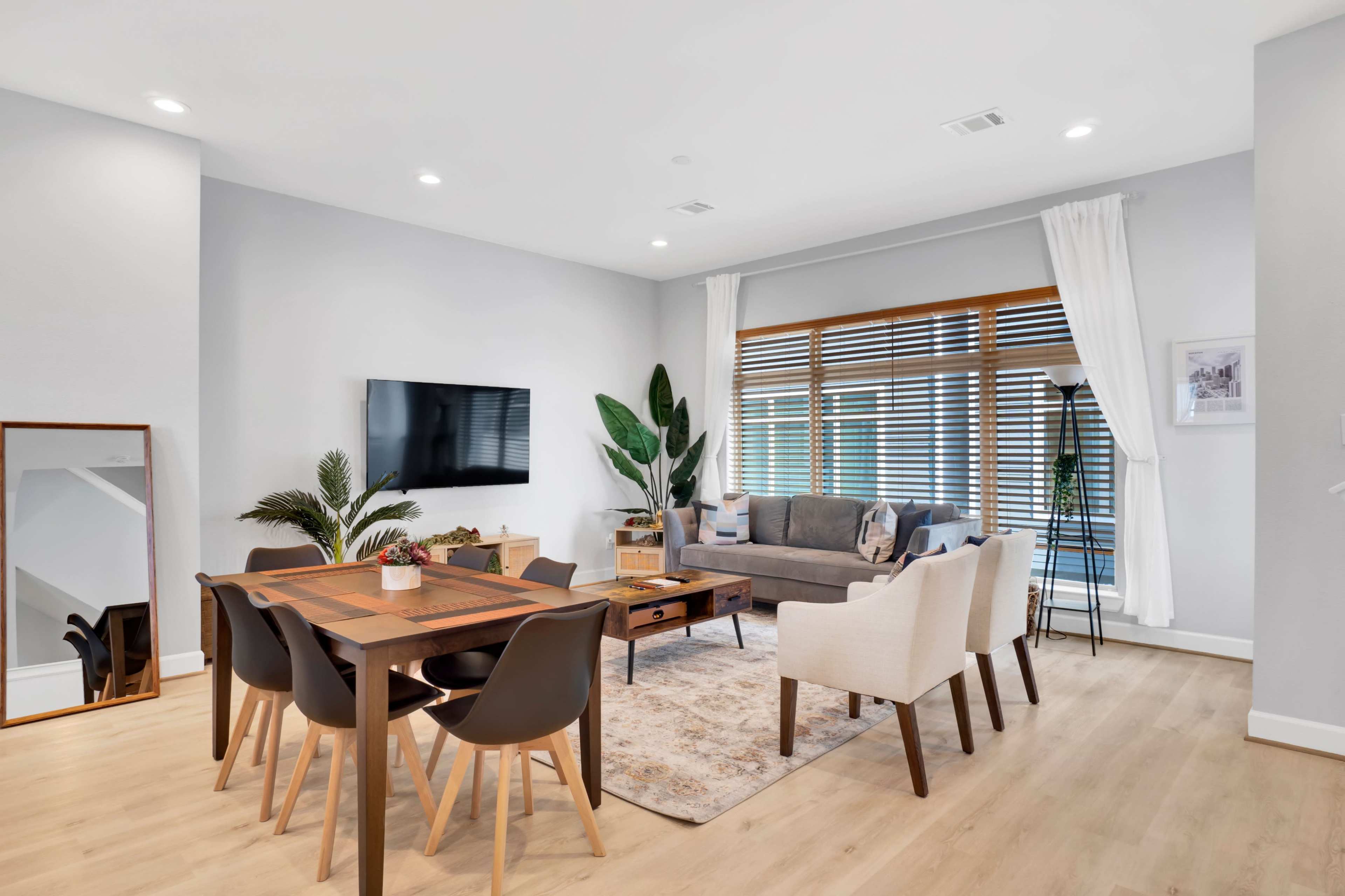 A modern living and dining area features a dining table with chairs, a sofa, and large windows with blinds, alongside a television mounted on the wall.