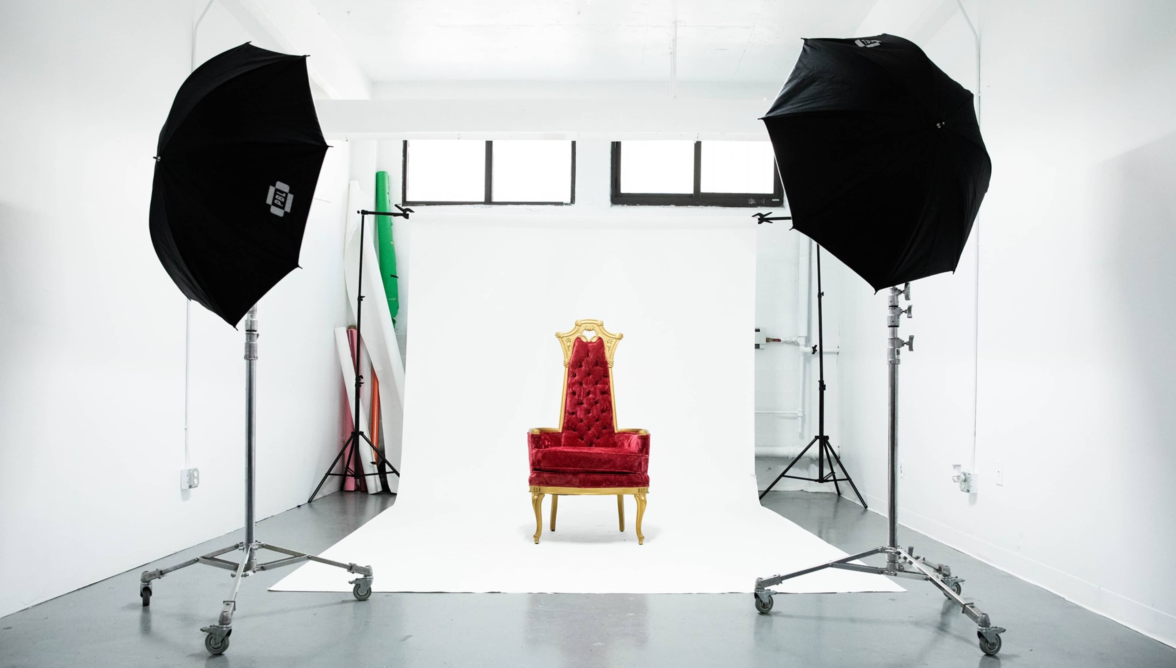 A red velvet throne is positioned on a white backdrop, flanked by two large black umbrella lights in a photography studio.