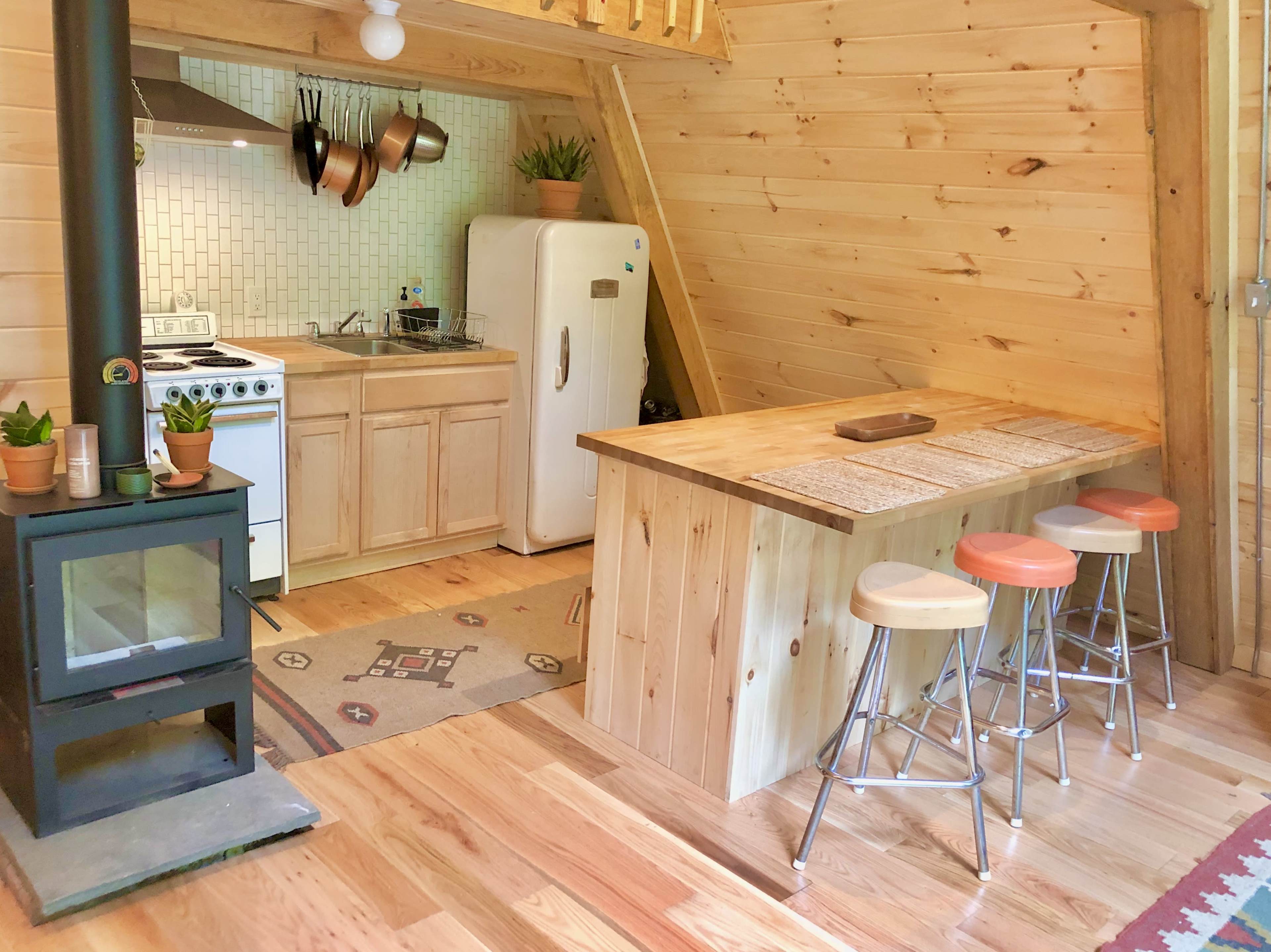 A cozy kitchen area features wooden cabinets, a white refrigerator, and a stove, with a wooden bar and stools beside a small wood-burning stove.