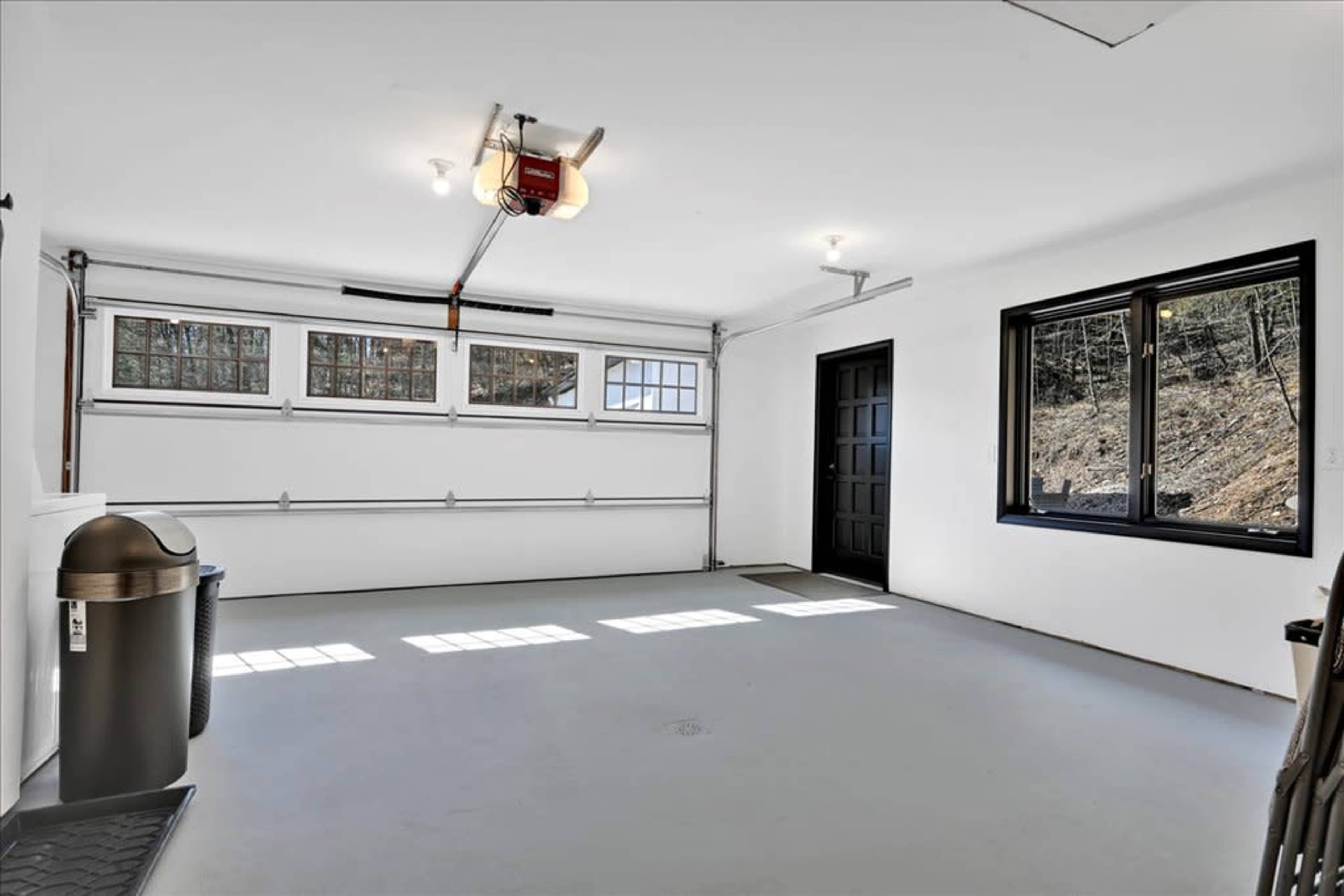 A spacious, empty garage with a large overhead door, a side entrance door, a window, and a trash can.
