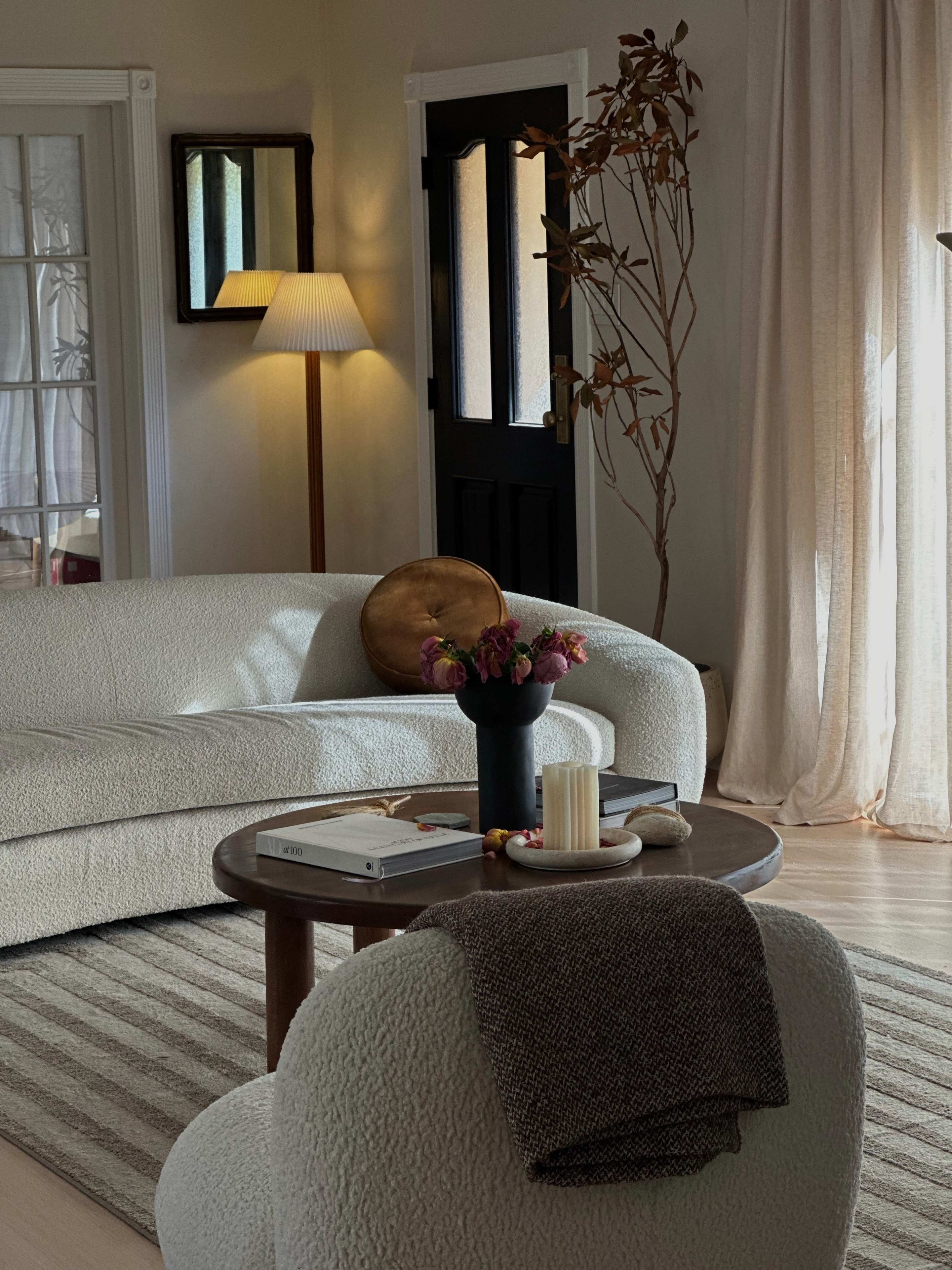 A cozy living room featuring a white, textured sofa, a round wooden table with a vase of flowers, and a tall lamp beside a dark door.