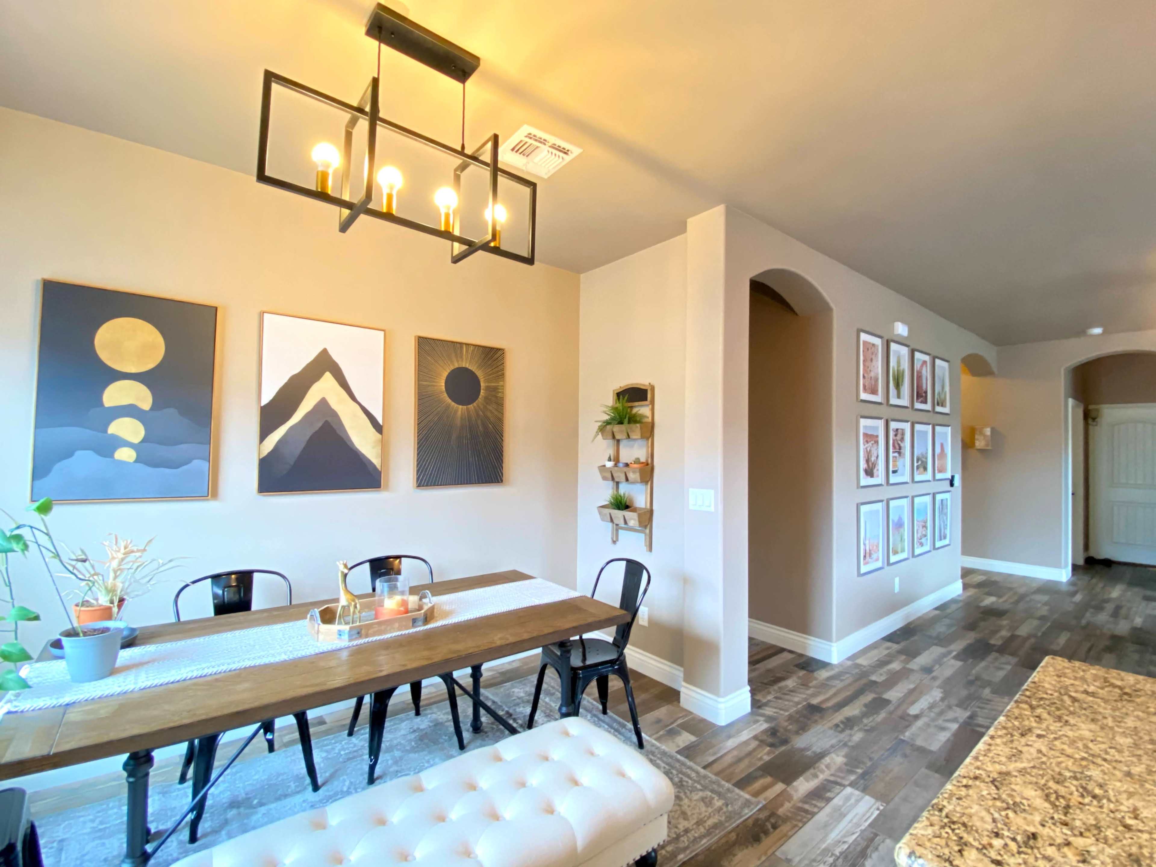 A dining area features a wooden table with black chairs, surrounded by framed artwork on the walls and a light fixture overhead.