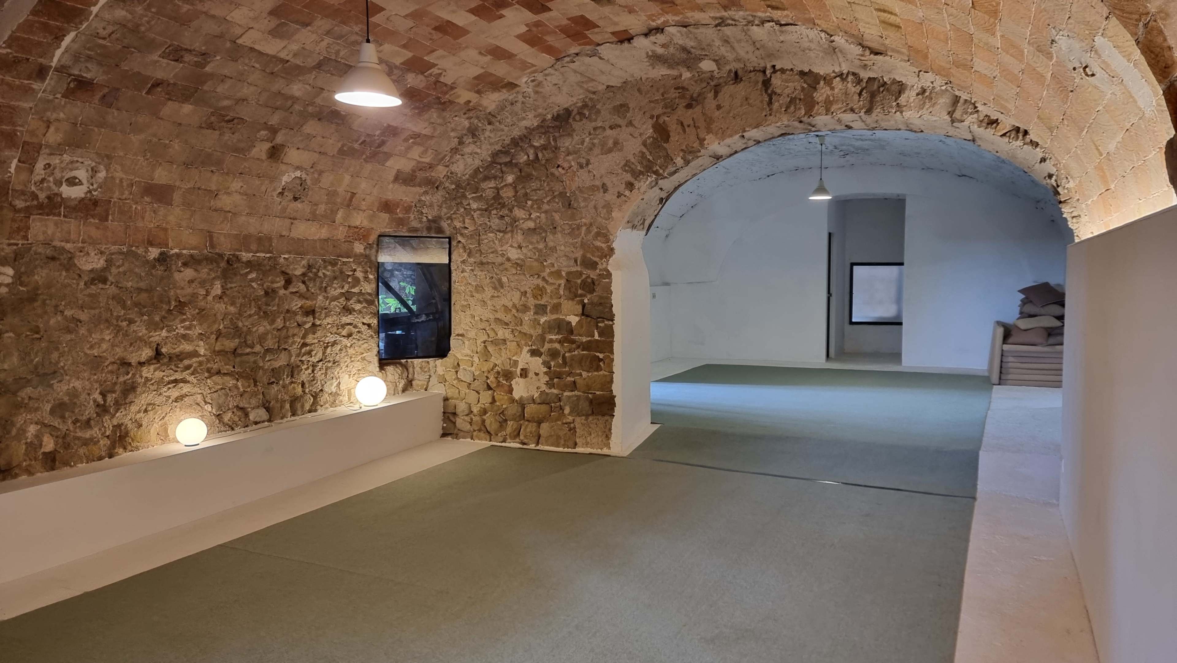 The image shows an interior space with arched brick ceilings, a stone wall on one side, and a carpeted floor illuminated by overhead lights.