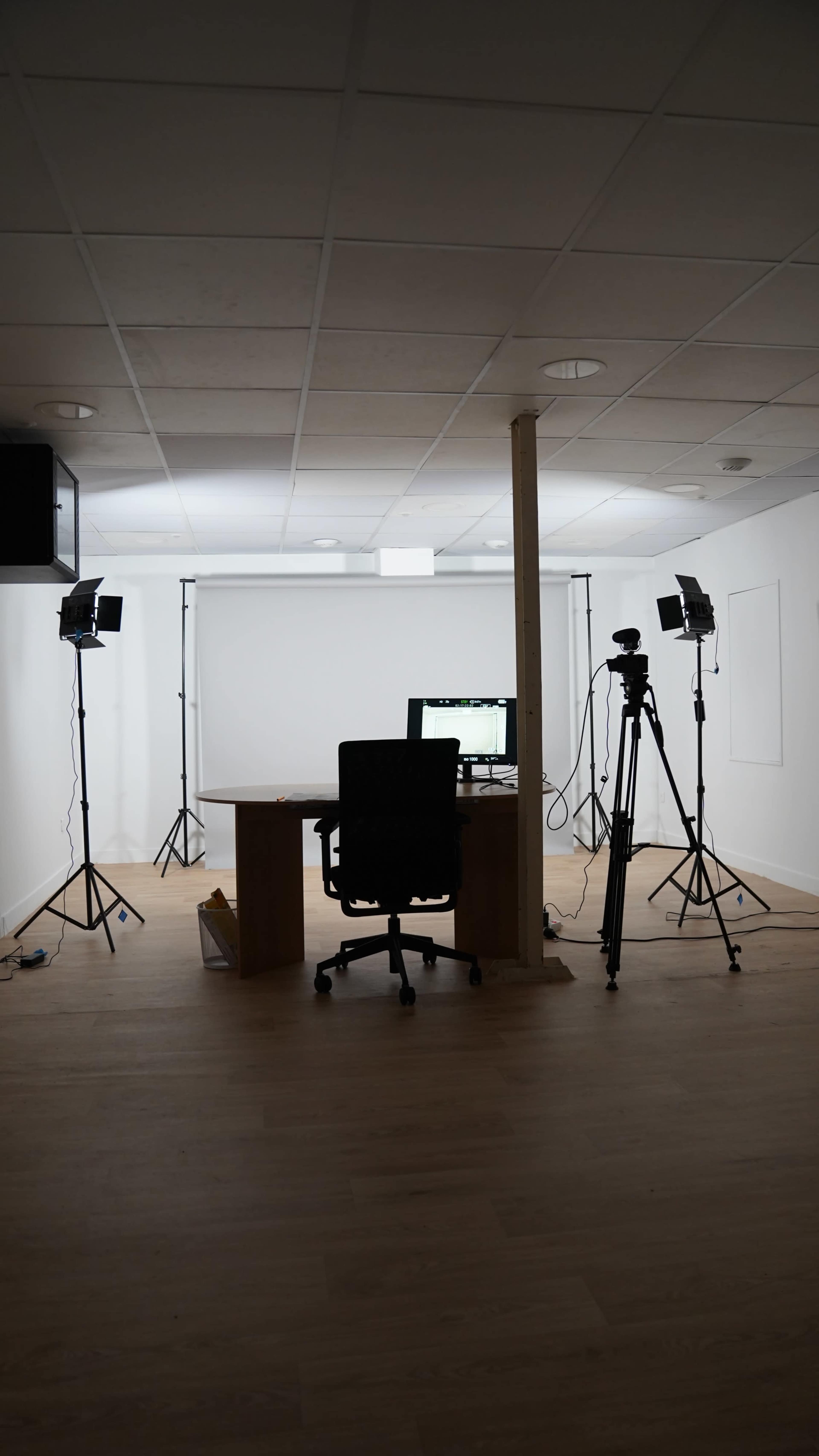 A minimalist office space features a desk with a computer, an office chair, and two lighting setups on tripods, accompanied by a backdrop for recording.