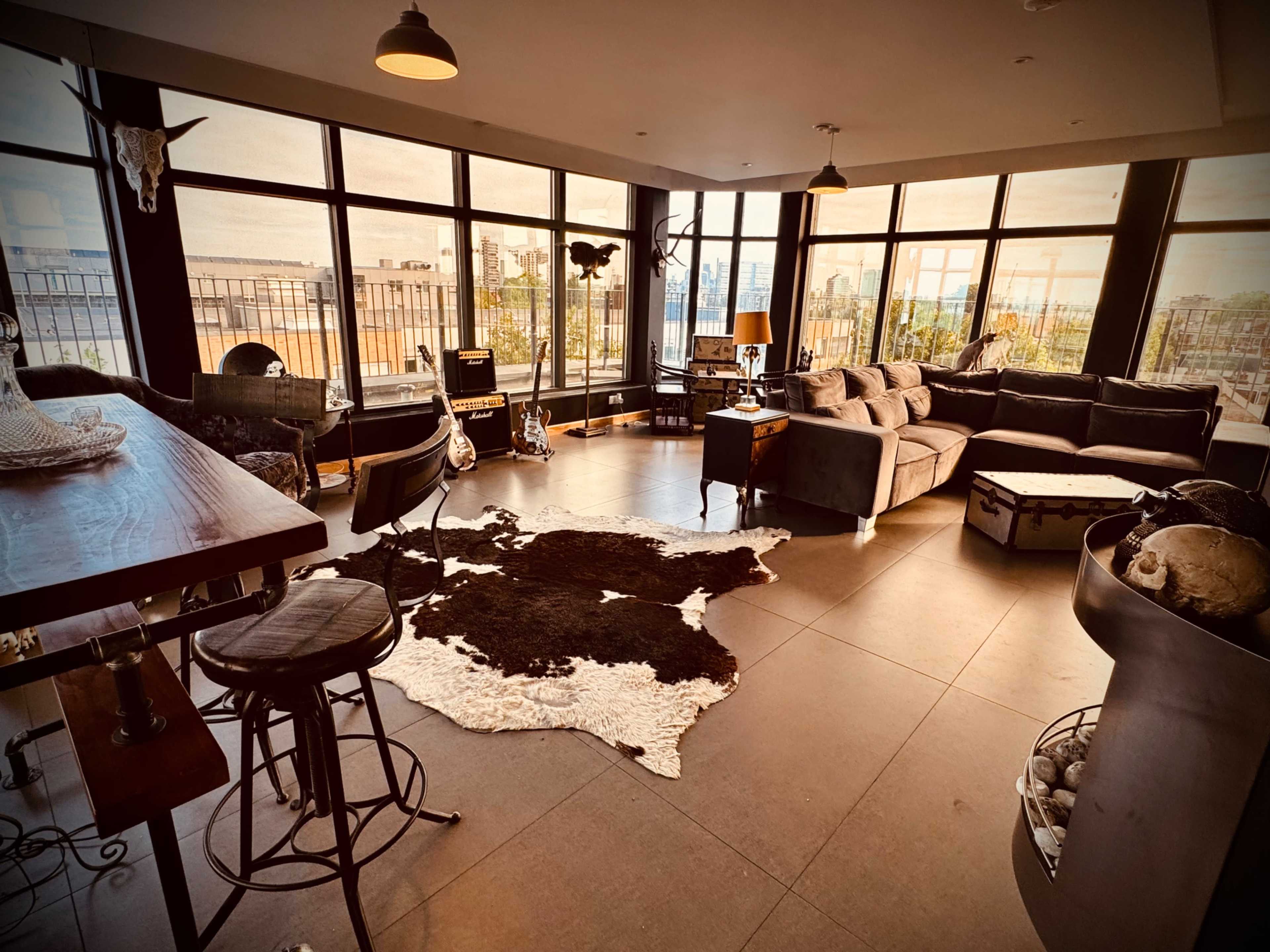 The image shows a modern, spacious living room with large windows, a sectional sofa, and a decorative cowhide rug on the floor.