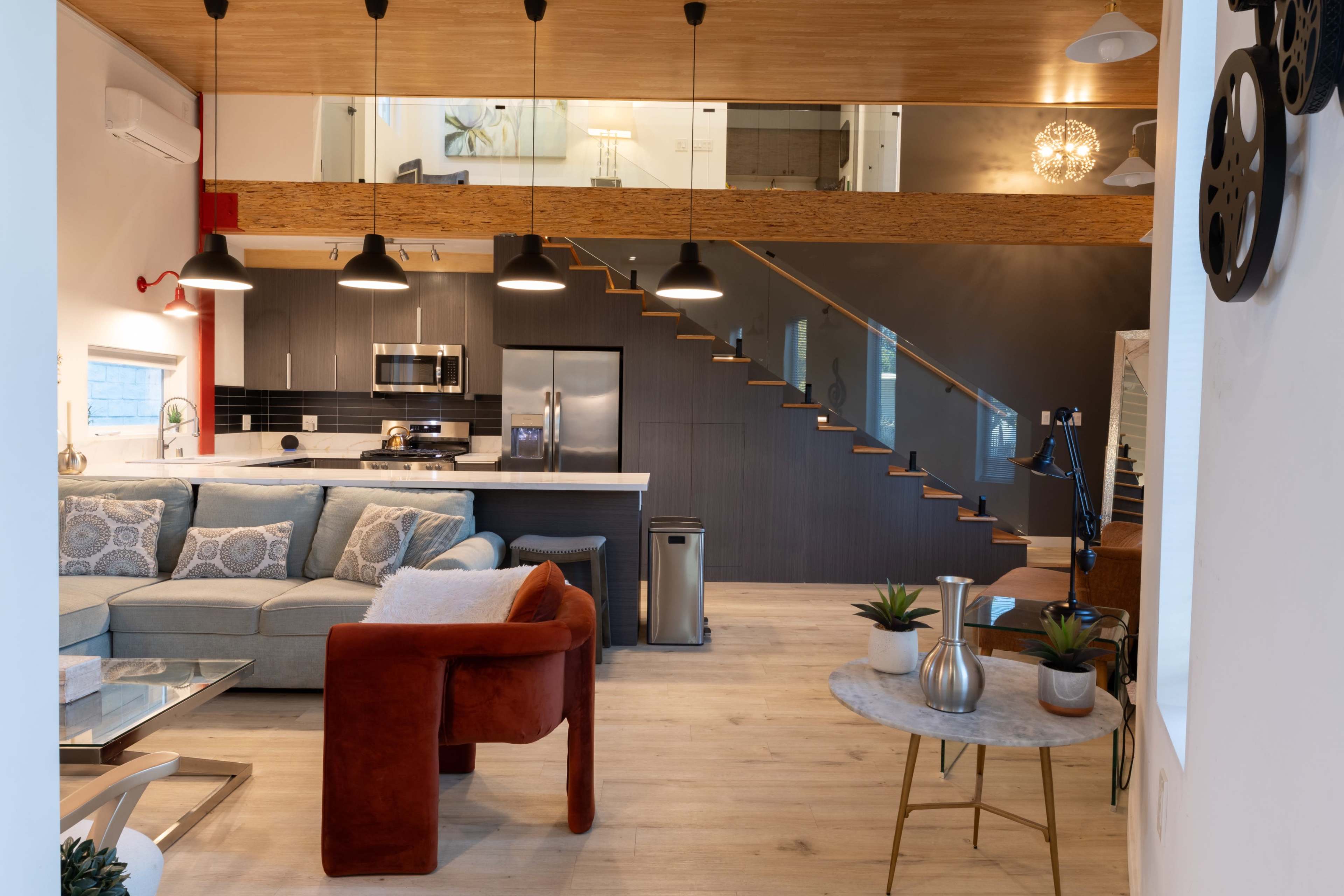 The image shows a modern open-concept living area featuring a sofa, an armchair, a kitchen with stainless steel appliances, and a staircase leading to an upper level.