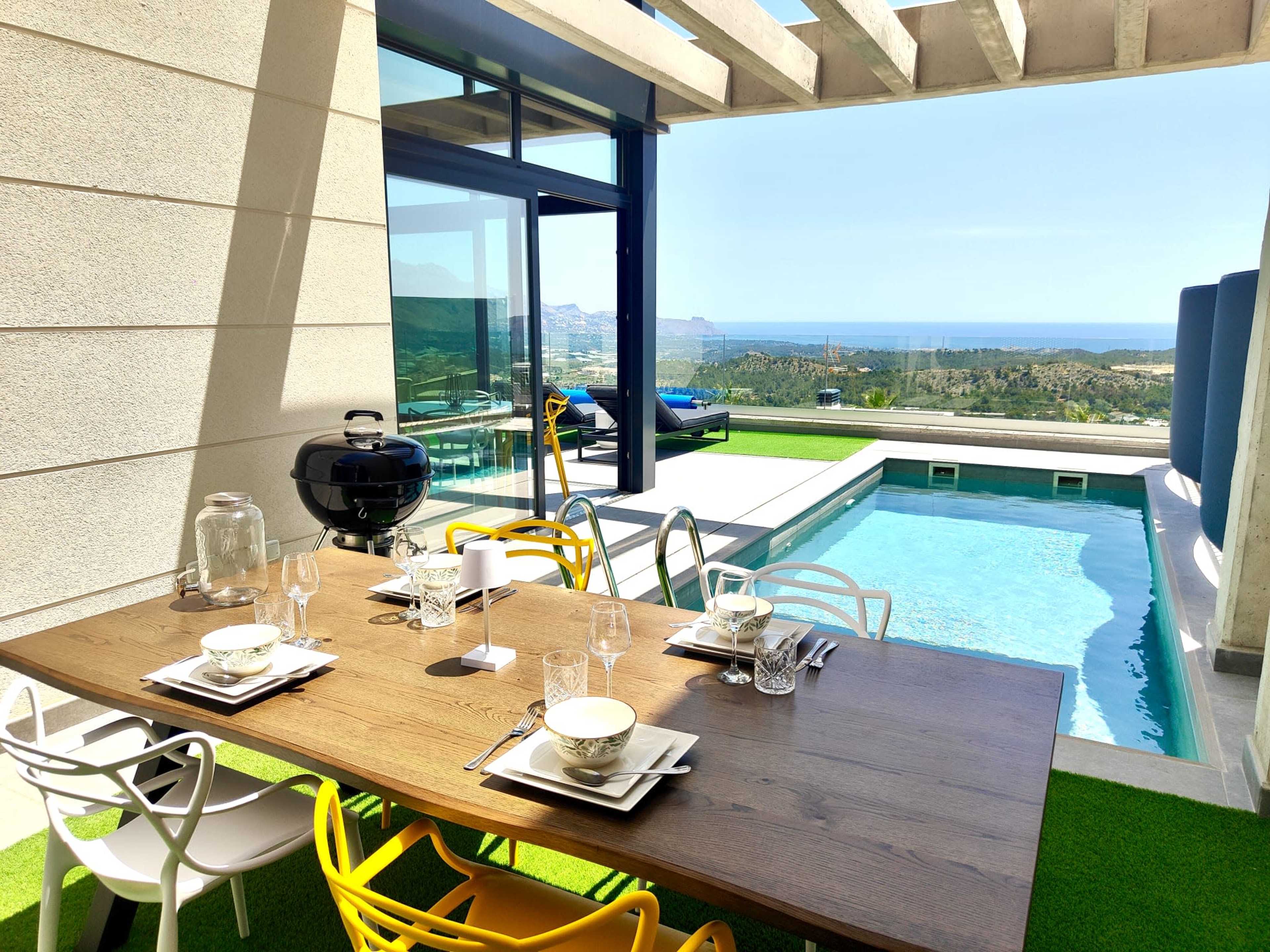 The image shows a modern outdoor dining area with a wooden table set for a meal, overlooking a pool and a scenic view of the coastline.