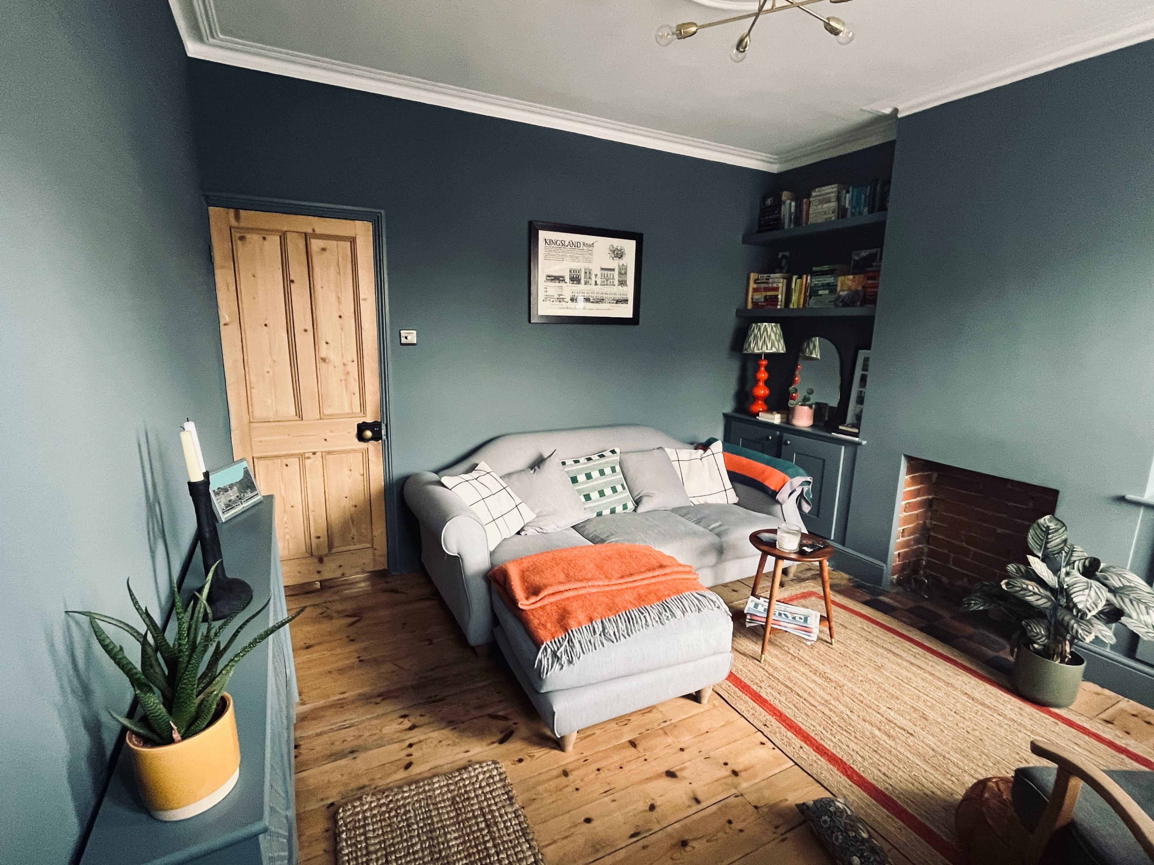 A cozy living room features a light gray sofa, wooden door, bookshelf, and a patterned rug, with a plant in the corner and a decorative orange throw.