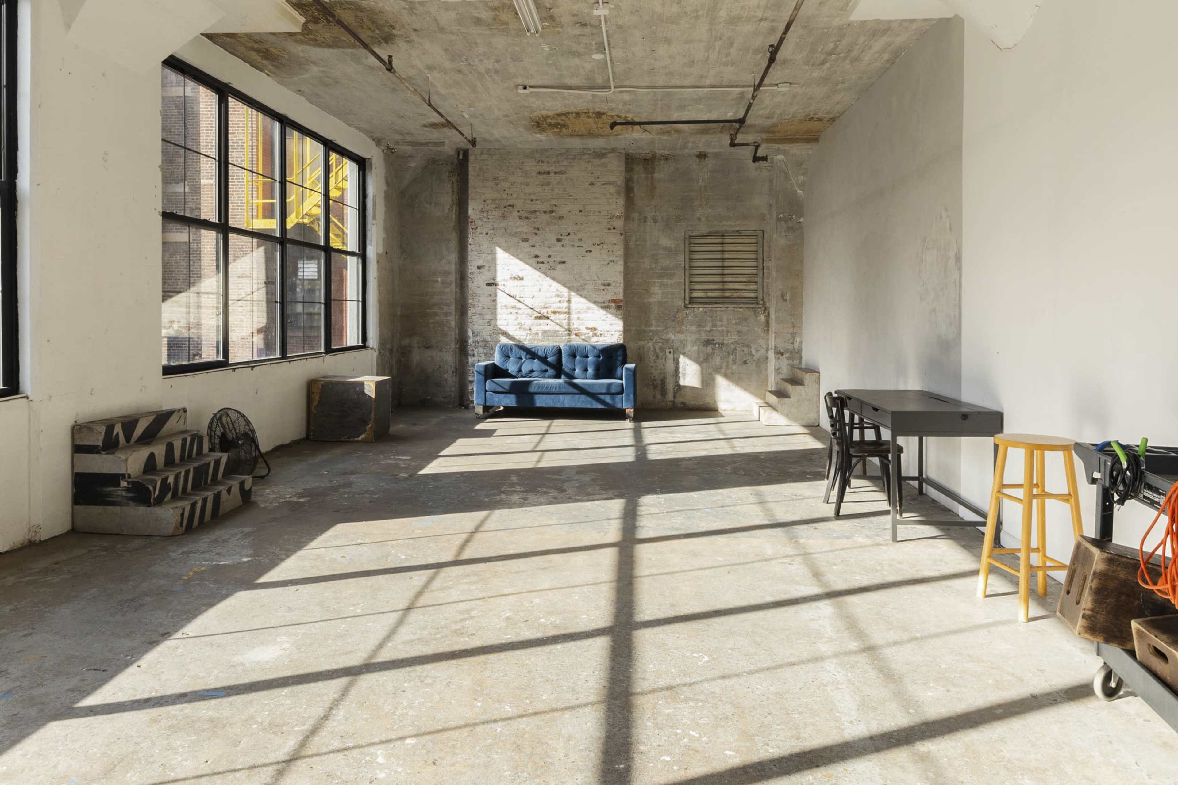 An empty industrial room features a blue couch, a table with stools, and large windows casting shadows on the concrete floor.